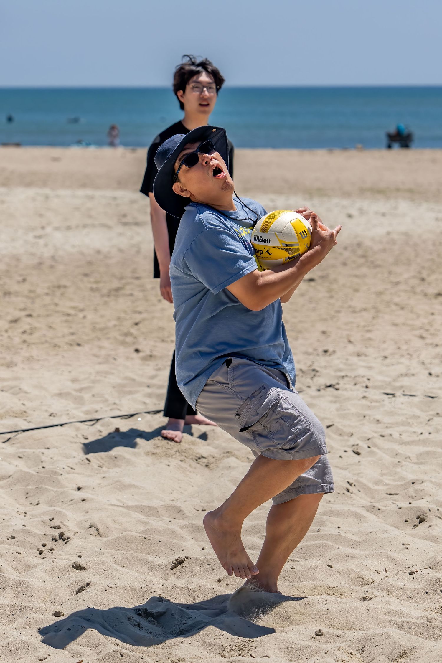Man in sunglasses bumping a yellow volleyball on a beach with another player watching behind him.