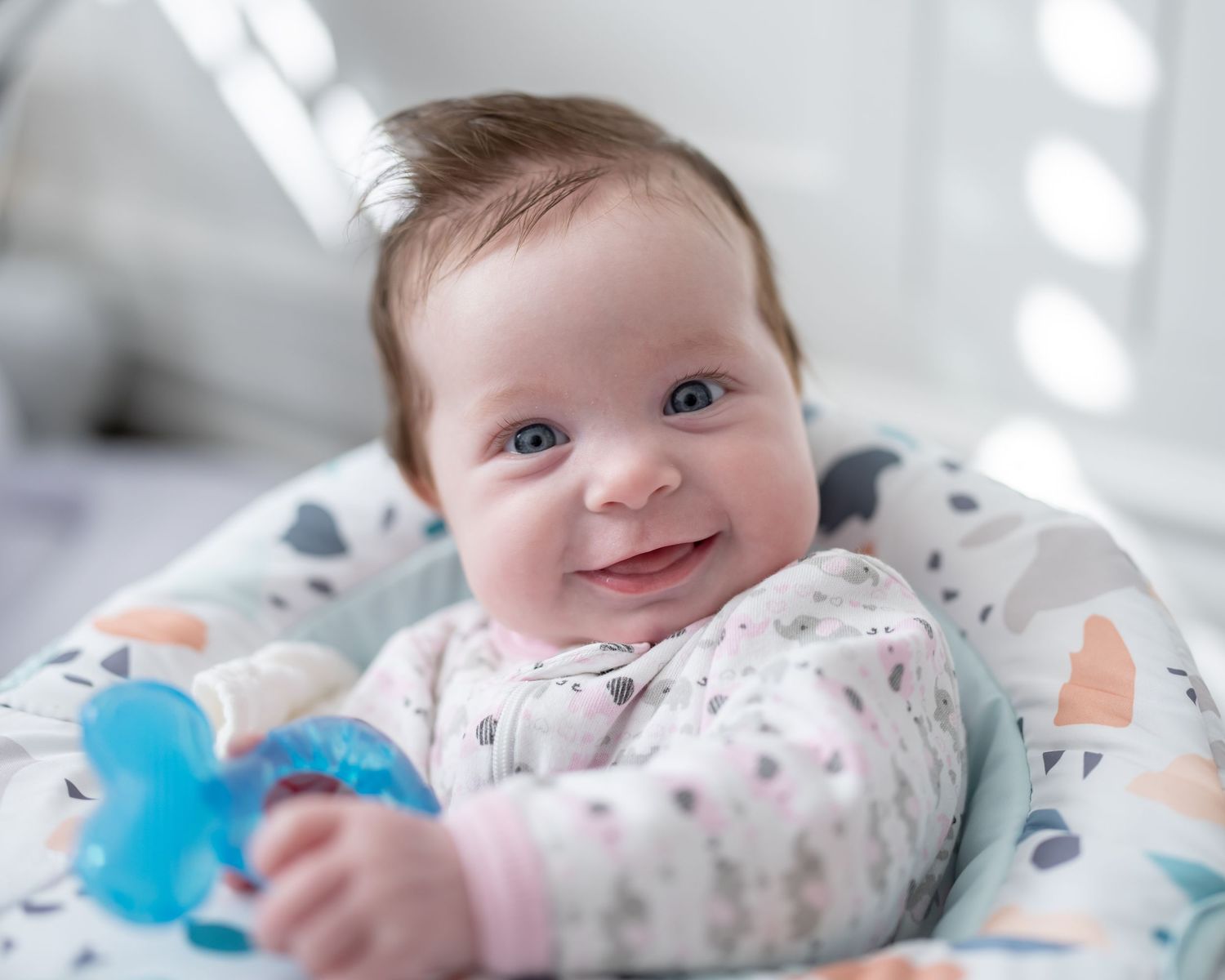Smiling baby with dark hair holding a blue pacifier, lying in a patterned infant seat.