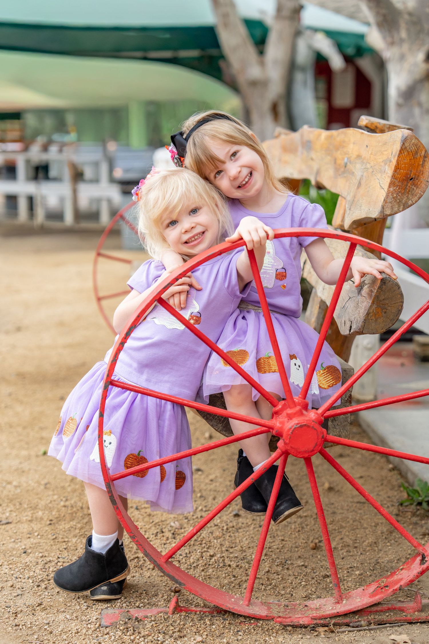 Two young girls in matching purple dresses pose by a red wagon wheel.