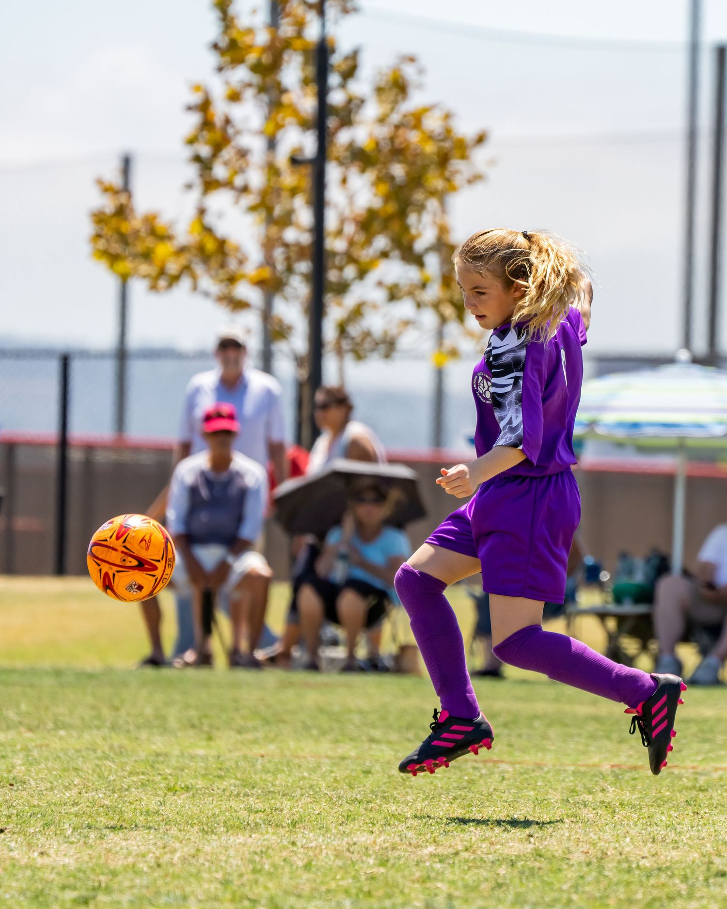 Young girl in purple soccer uniform kicking orange ball during youth soccer game outdoors.