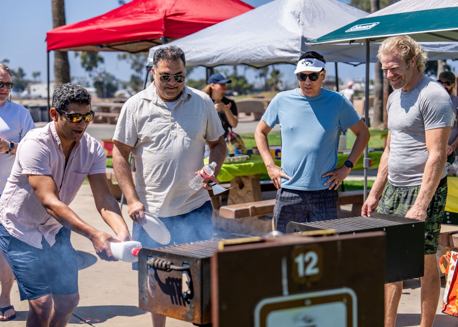 Men starting charcoal grill at outdoor BBQ event with red and white canopy tents nearby.