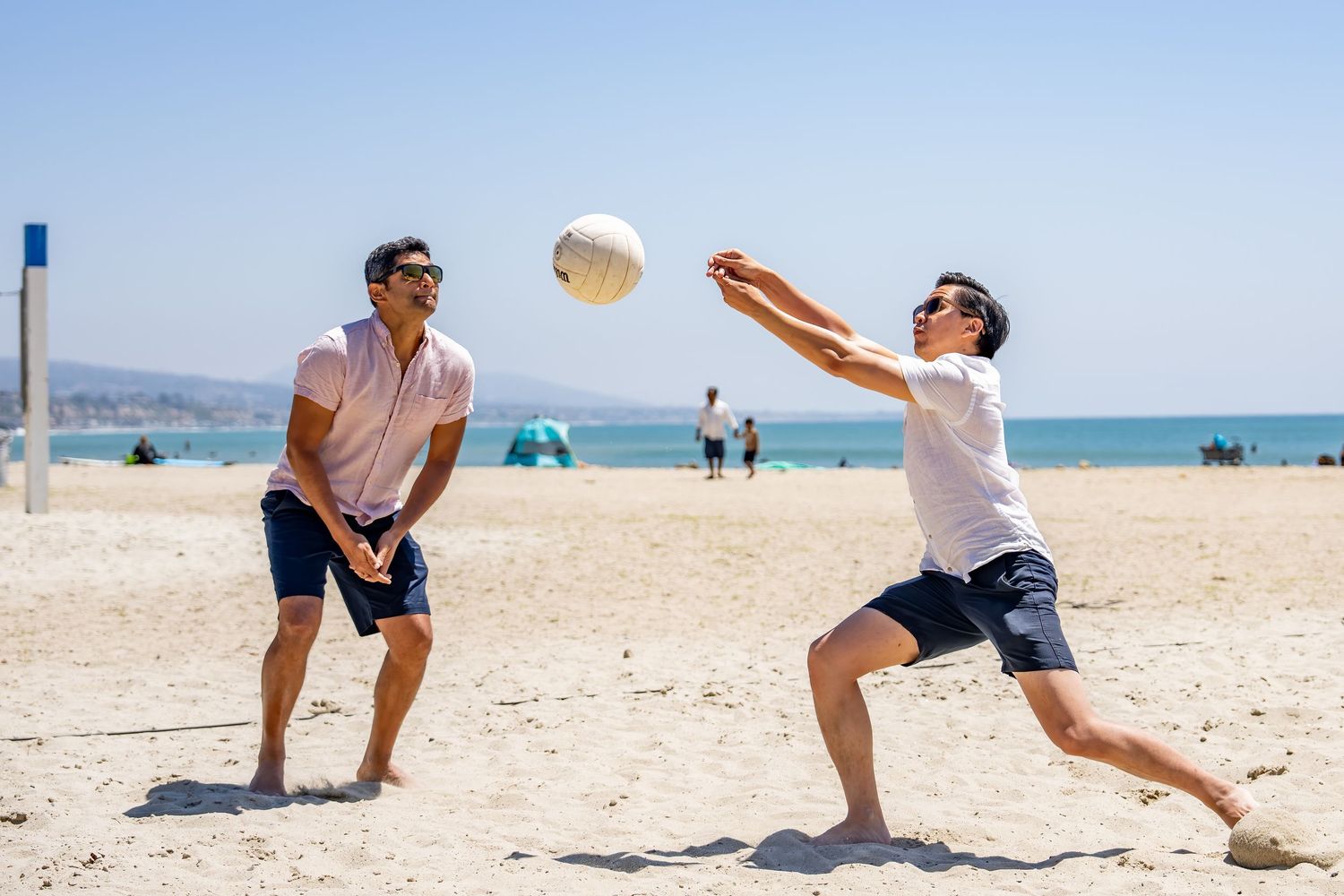 Two men playing beach volleyball on a sunny beach, one lunging to bump the white ball.
