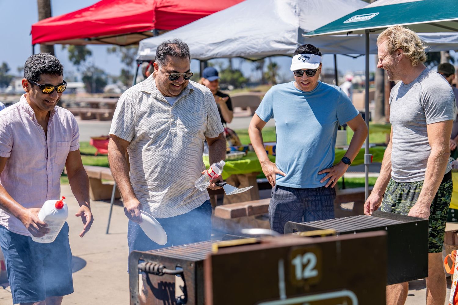Four men gathered around charcoal grill at outdoor BBQ cookout under colorful canopy tents.