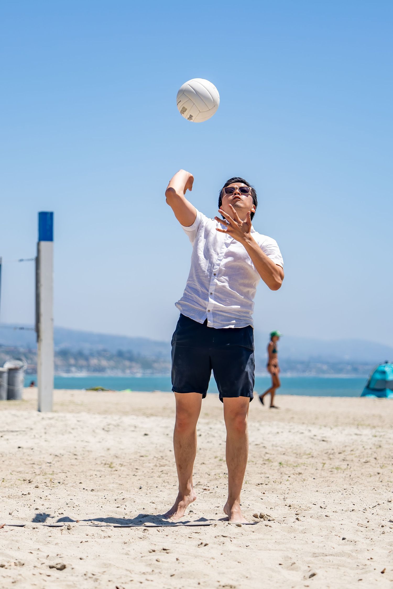 Man in white shirt and sunglasses serving a volleyball on a sunny beach.