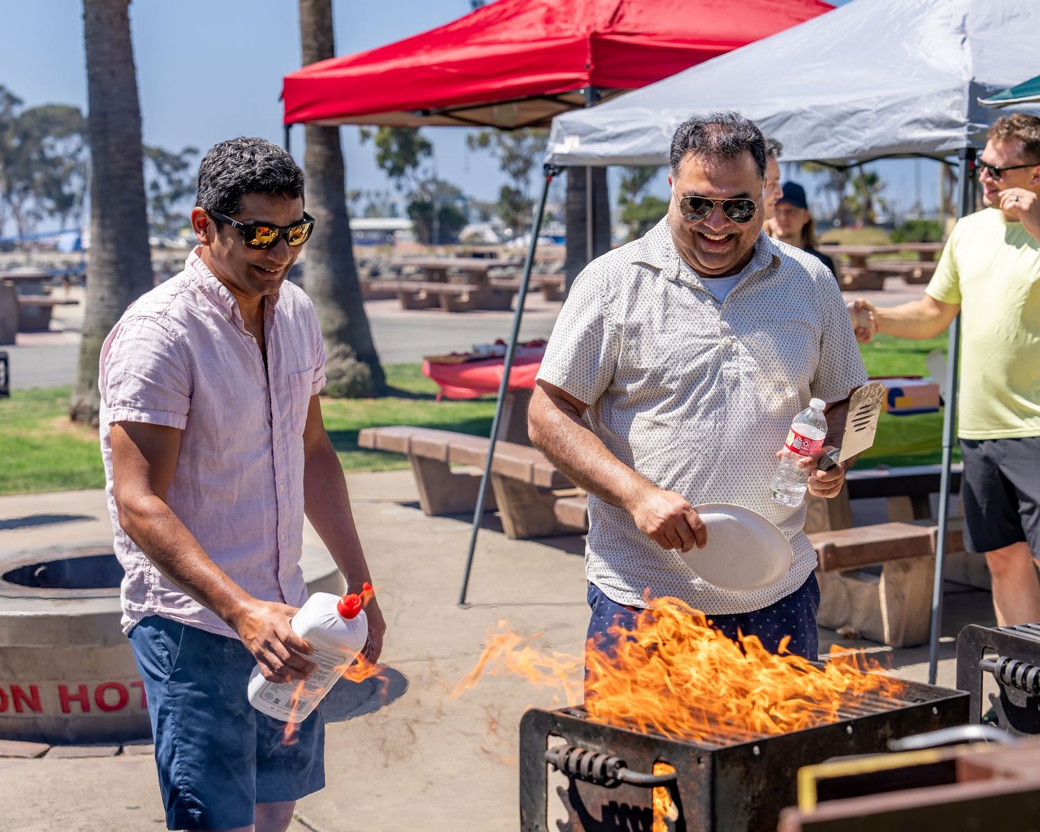 Two men laughing while tending to a large open flame on an outdoor grill at a park.