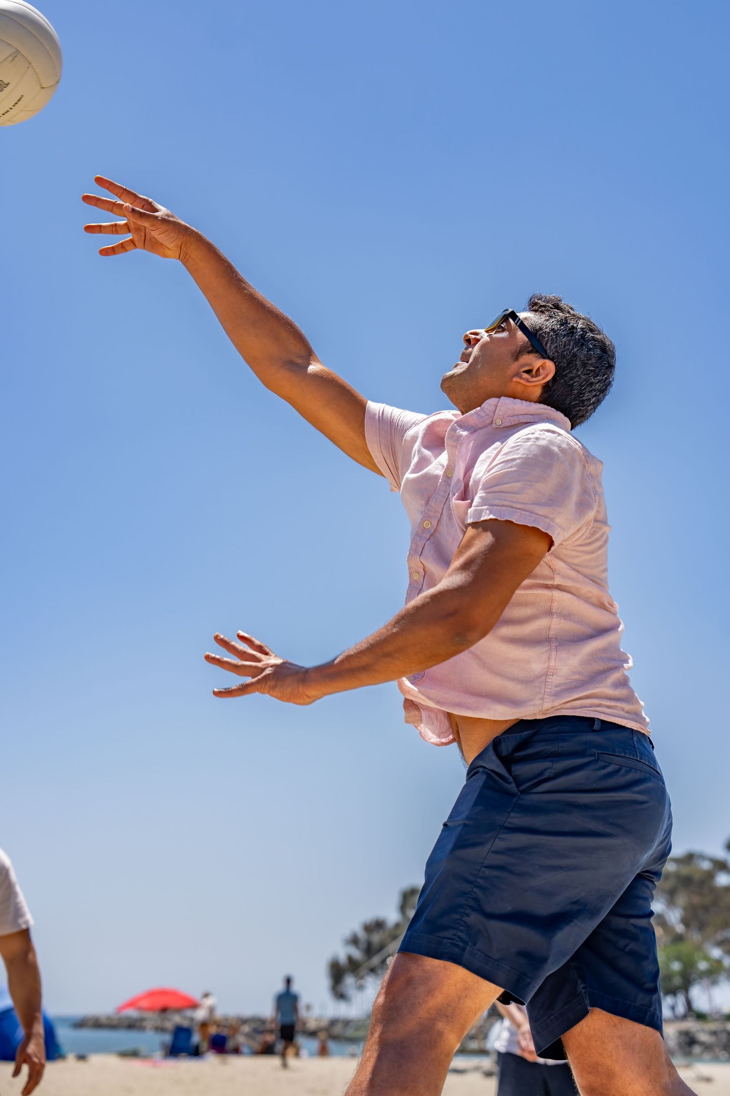 Man in pink shirt reaches up to hit volleyball on sunny beach.