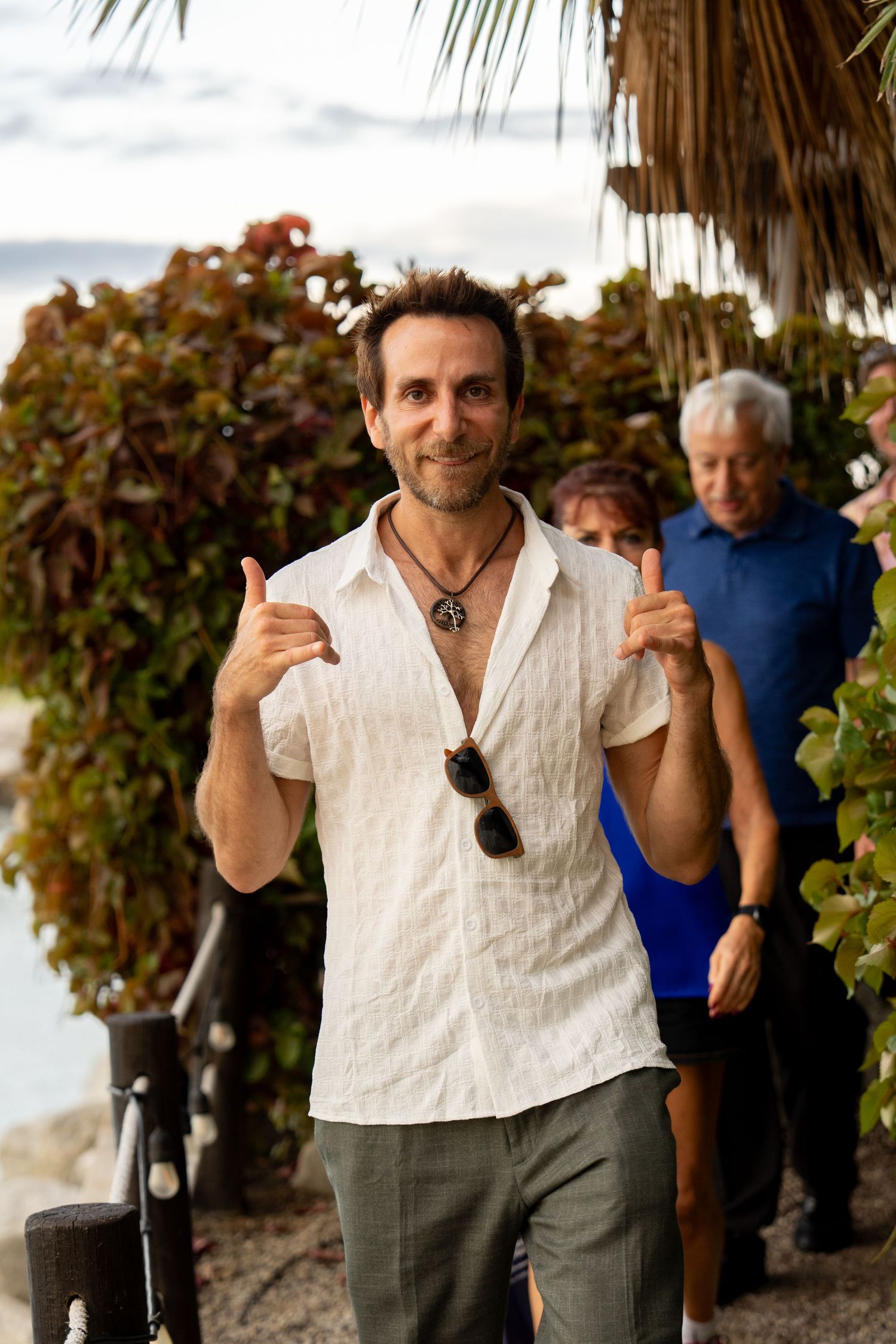 Happy man in white shirt giving thumbs up at a tropical outdoor beach venue.