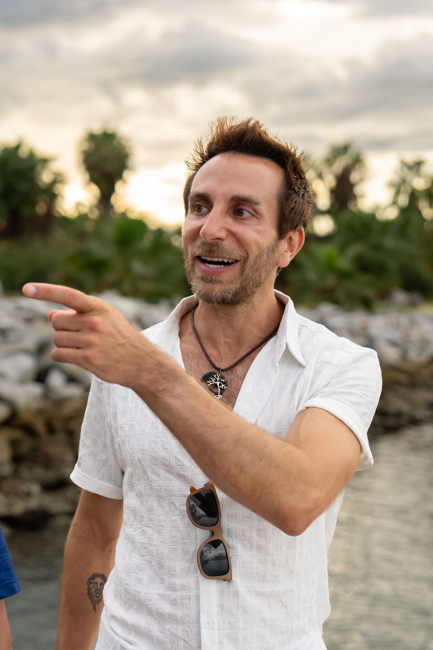Man in white linen shirt pointing and smiling at a tropical waterfront sunset.