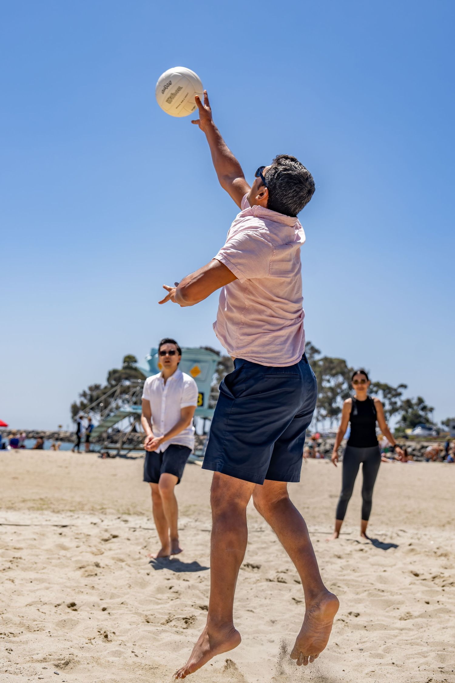 Man in pink shirt jumps to set volleyball on crowded sunny beach.