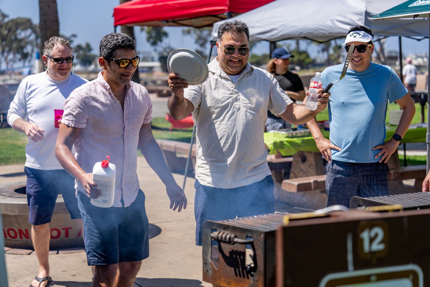 Group of men grilling food at outdoor BBQ event near beach on sunny day.
