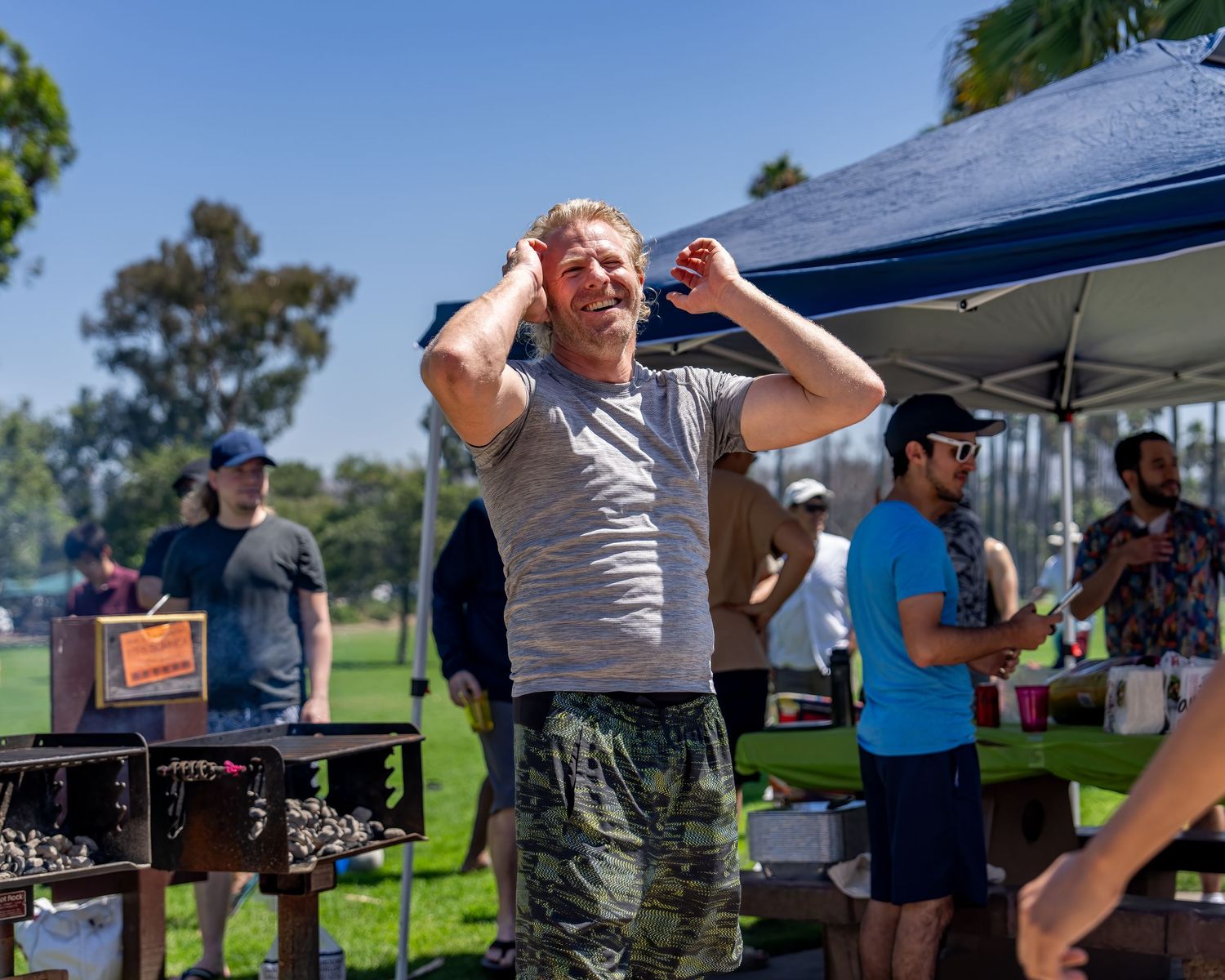 Blond man laughing with hands on his head near outdoor grills at a sunny park event.