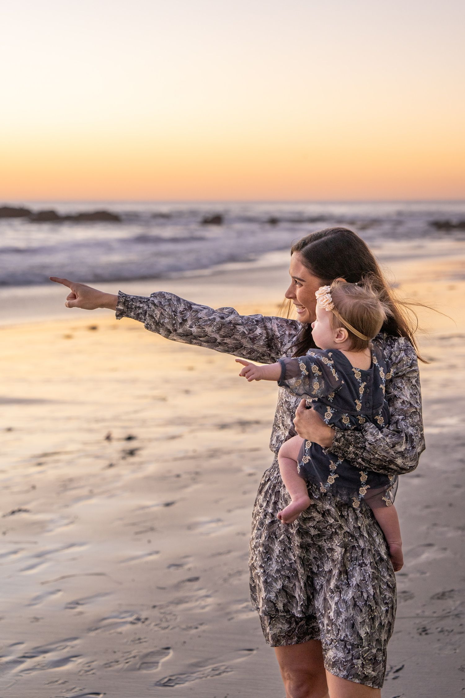 Mother holding baby pointing toward ocean horizon during golden sunset on sandy beach.