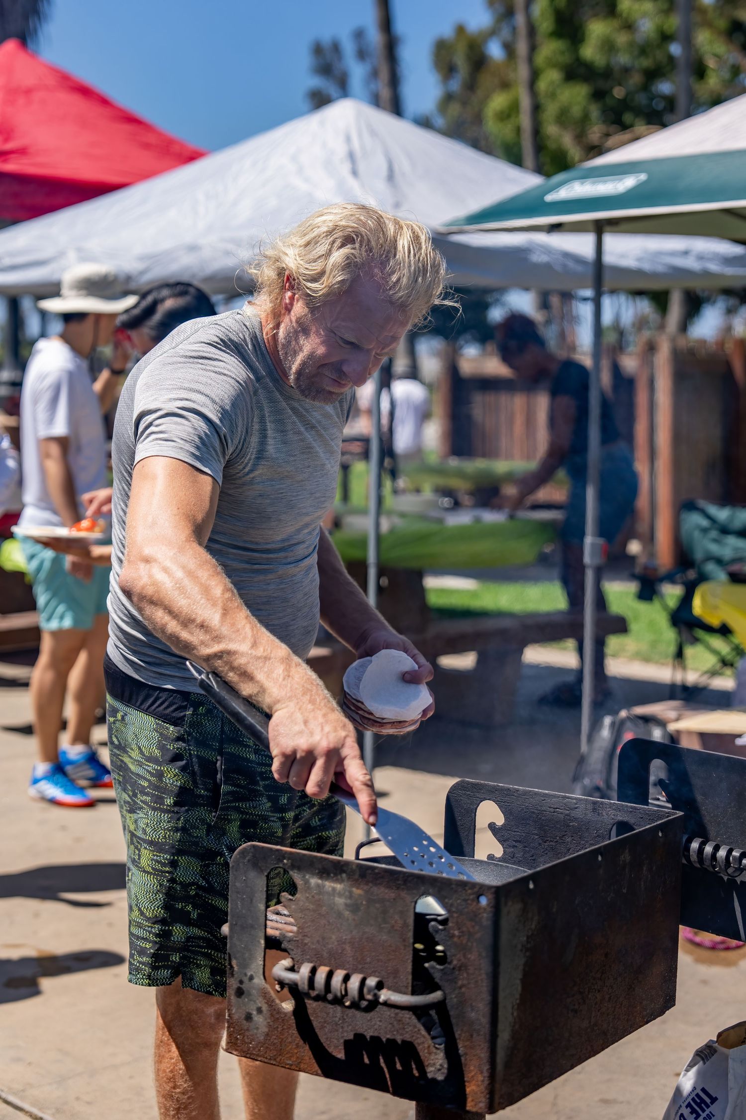 Older man with gray beard using a spatula to grill food at a sunny outdoor barbecue event.