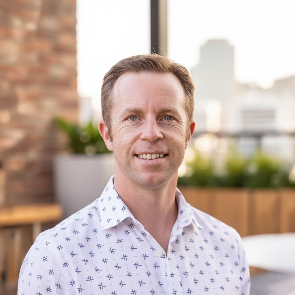 Cheerful man with auburn hair in a patterned white shirt posing on a sunny urban rooftop terrace.