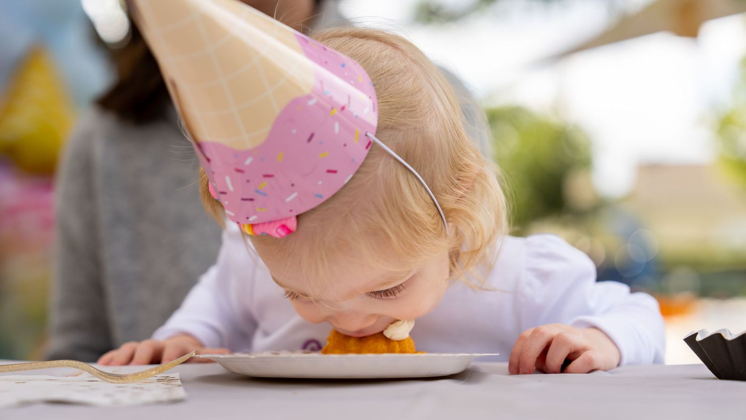 Blonde toddler wearing a pink ice cream party hat leaning down to eat from a plate outside.