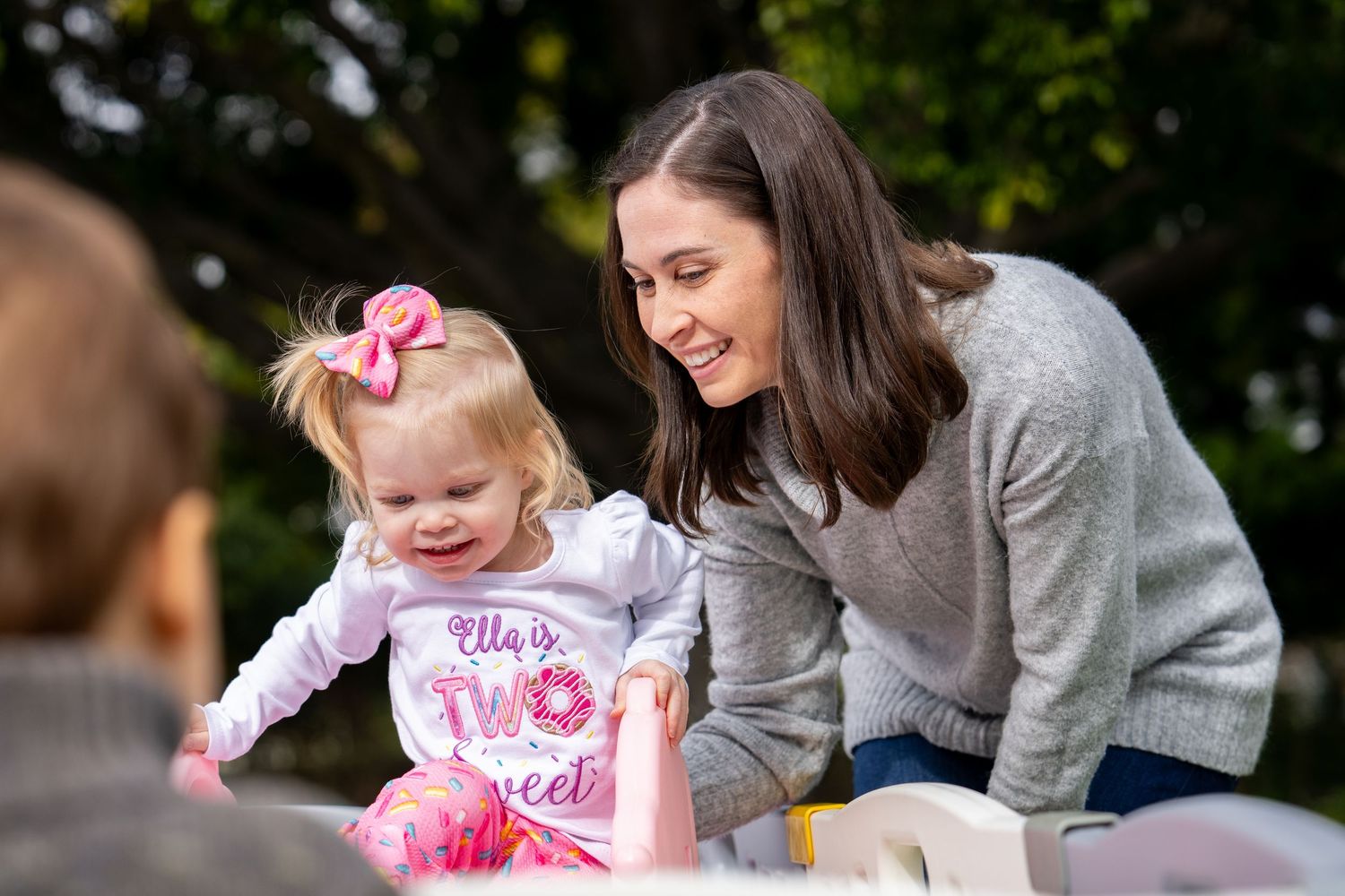 Smiling mother leaning over laughing baby girl in donut-themed birthday outfit outdoors.