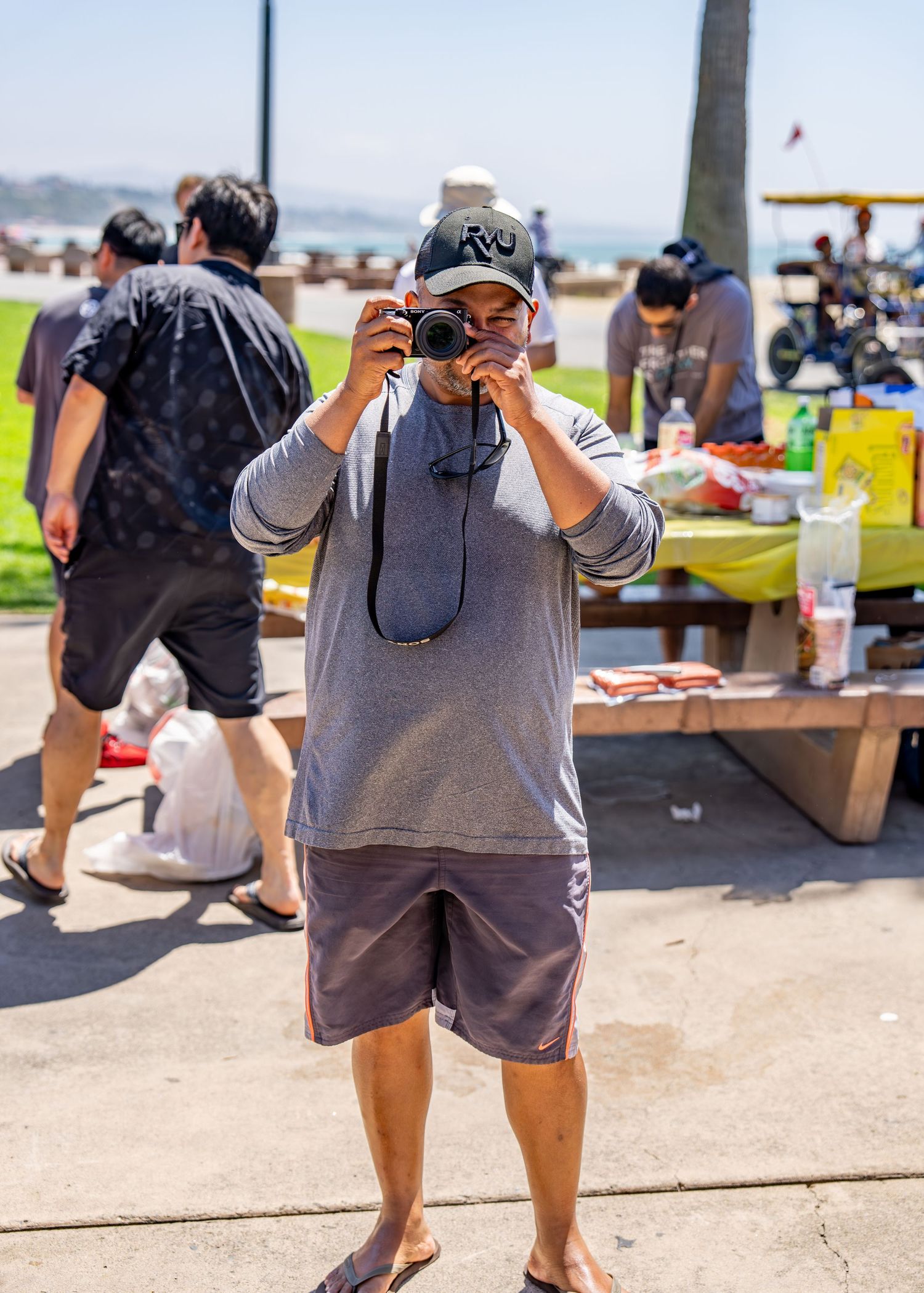 Man in gray hoodie and cap photographing with camera at outdoor beach park gathering.