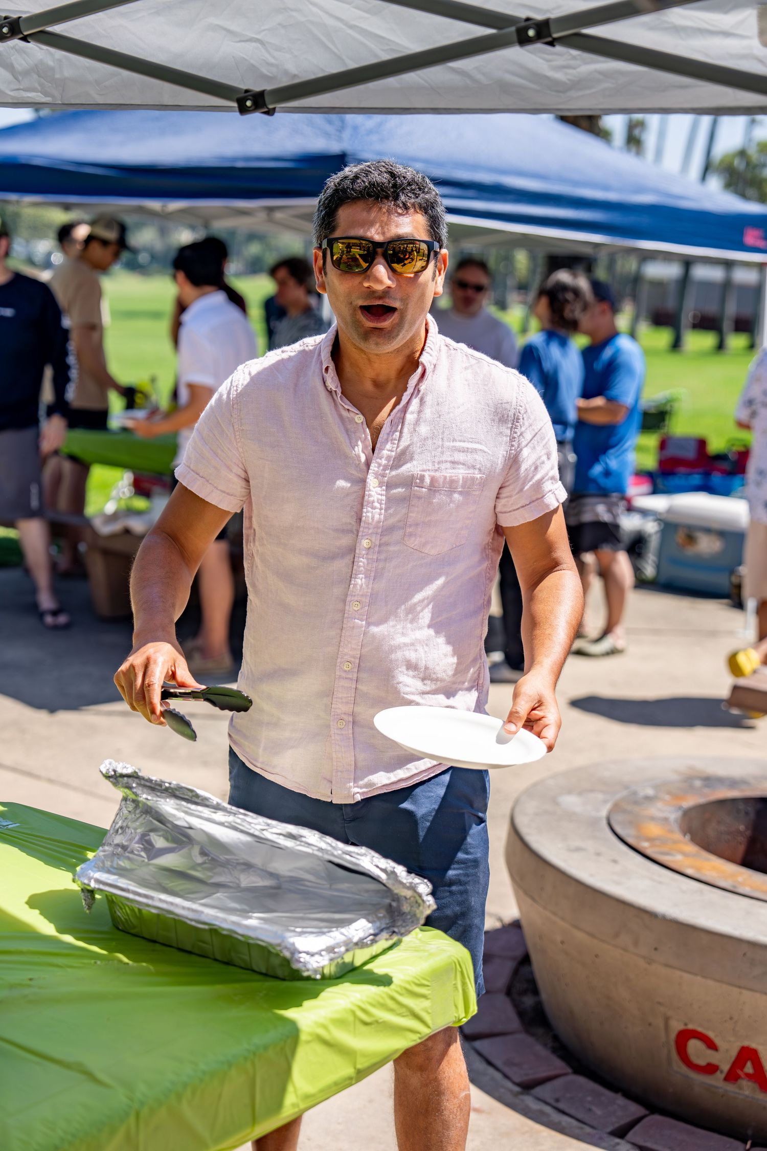 Man in pink shirt and sunglasses serving food from foil pan at outdoor BBQ event.
