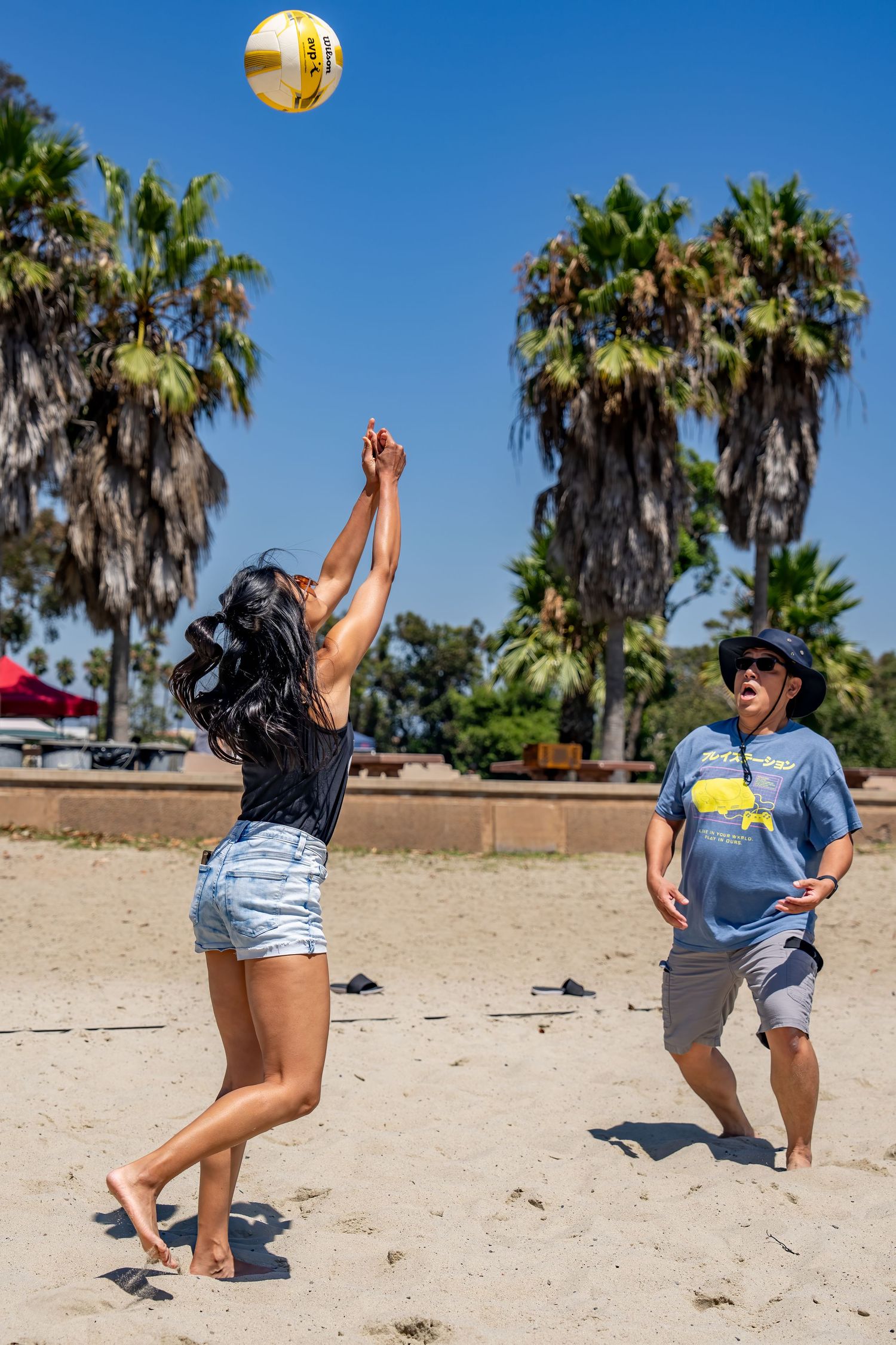 Two people play beach volleyball near palm trees on a sunny day.
