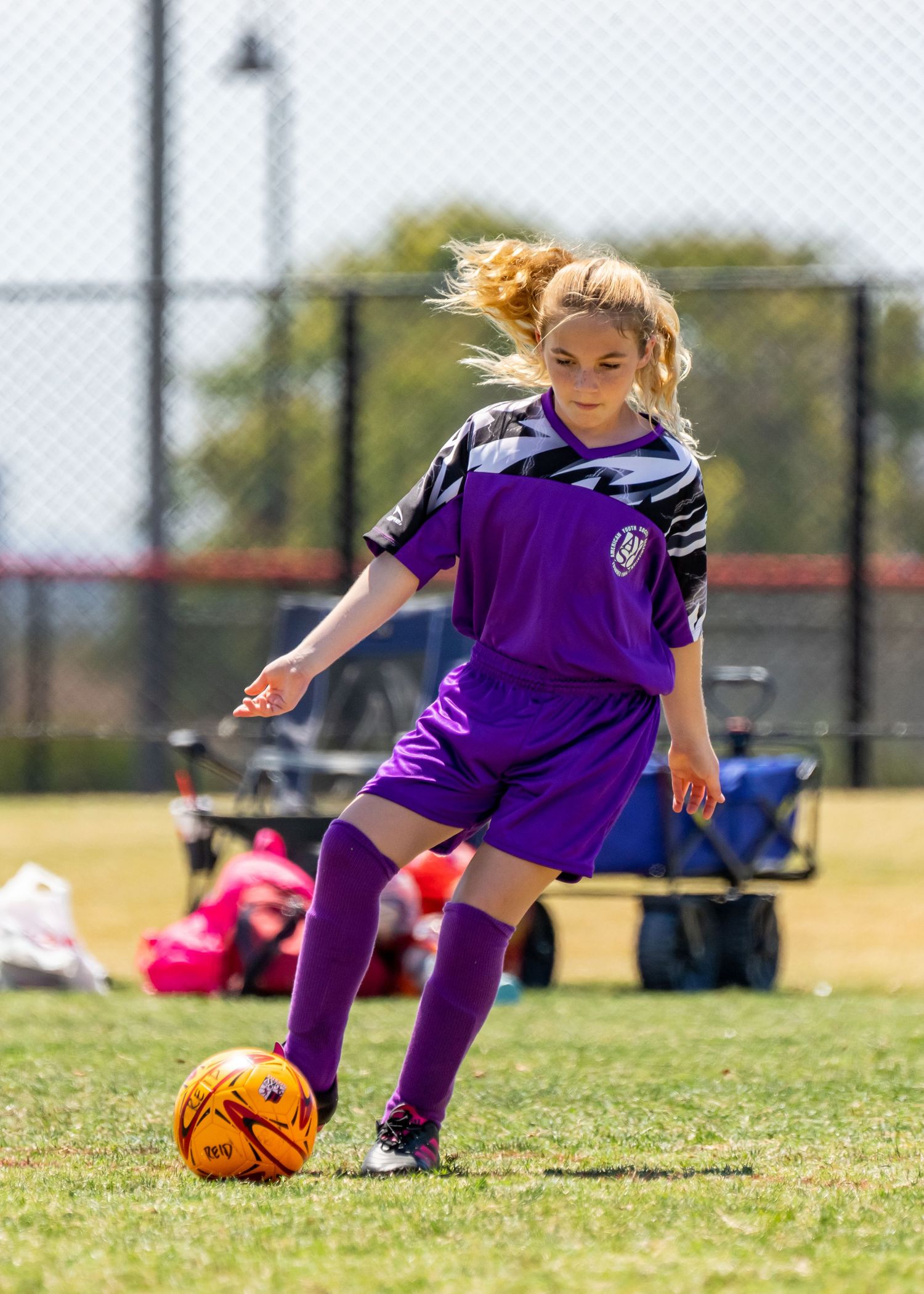 Blonde girl in purple soccer uniform controlling orange ball on sunny outdoor soccer field.