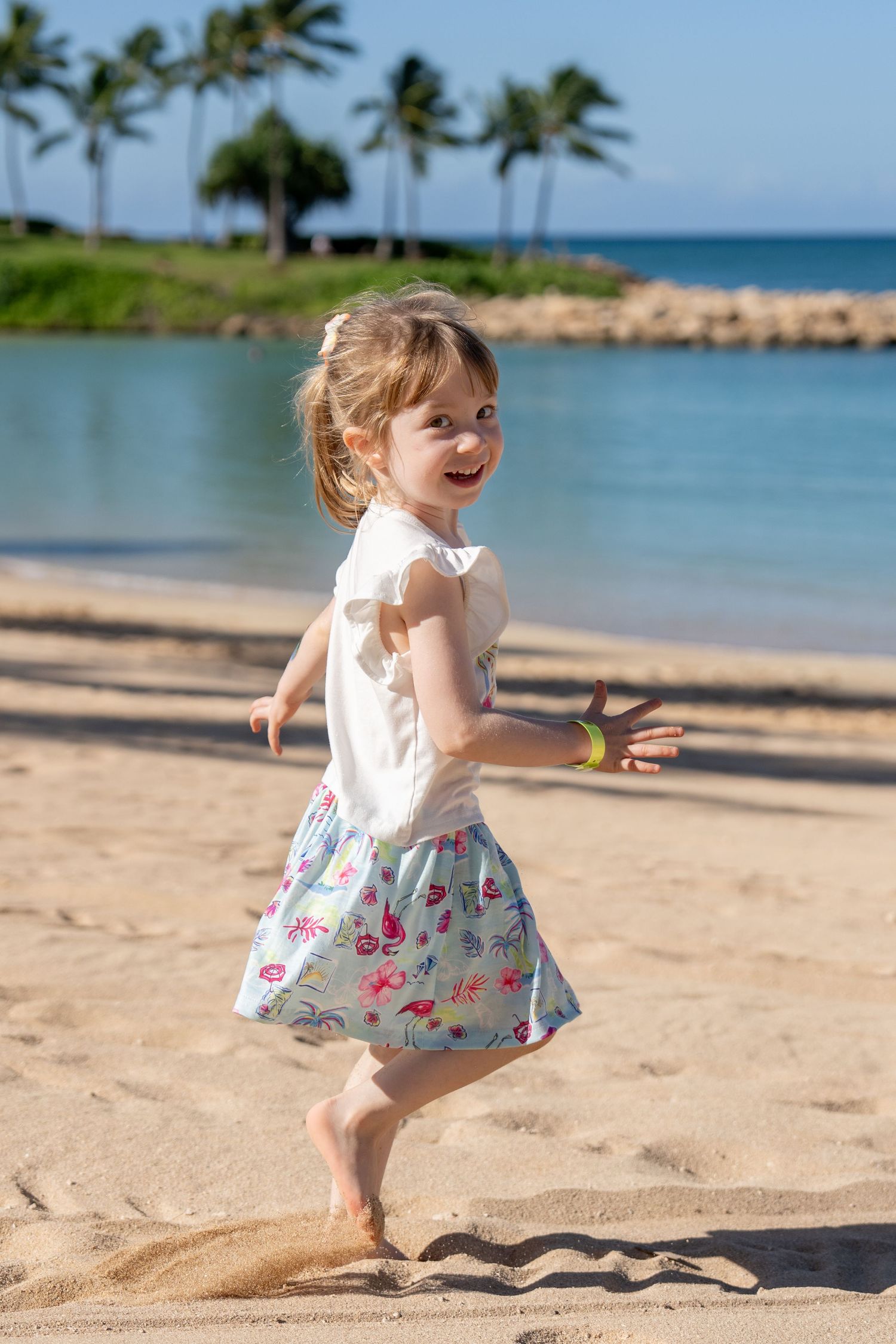 Happy toddler girl in skirt running barefoot on sandy tropical beach near calm water.