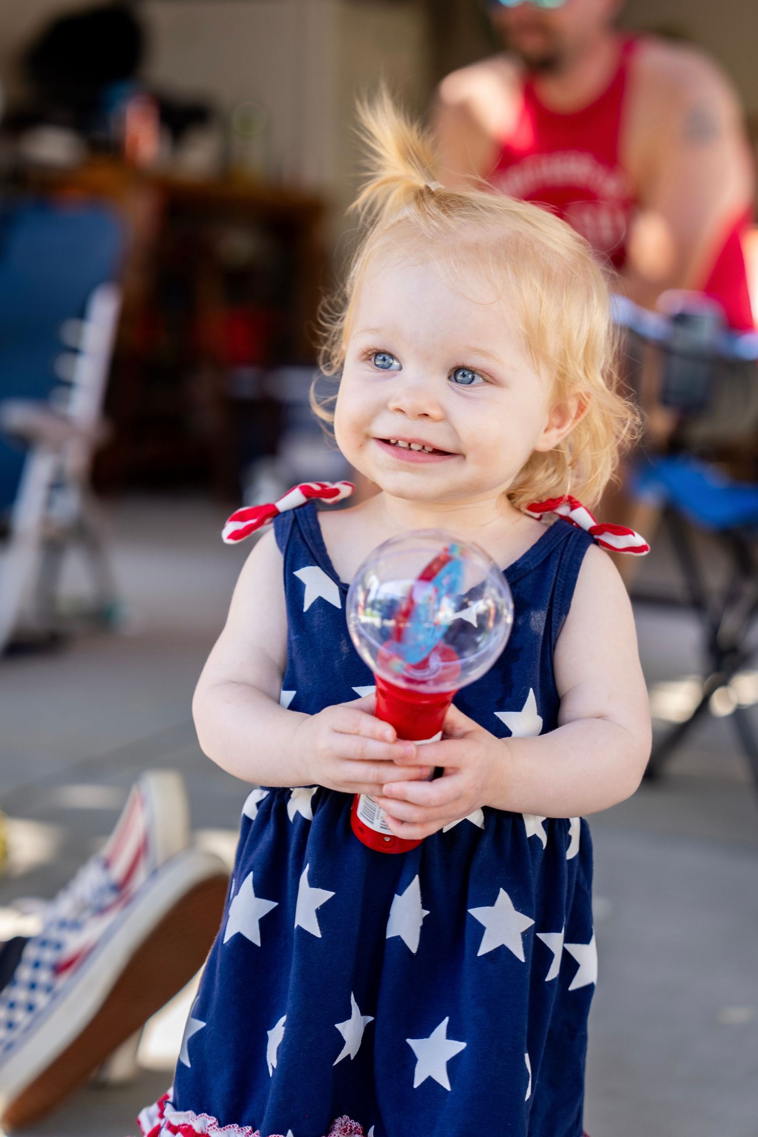 Happy blonde toddler girl in patriotic star dress holding a red white and blue maraca toy.