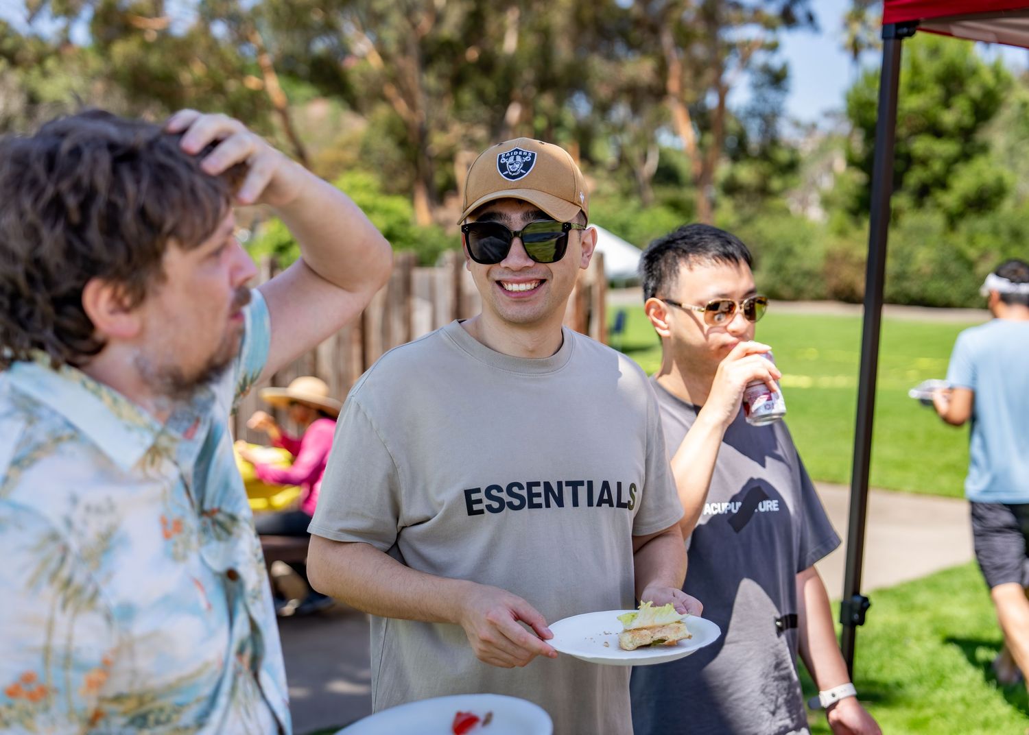 Young man in Essentials t-shirt smiling and holding a plate at a sunny outdoor social event.