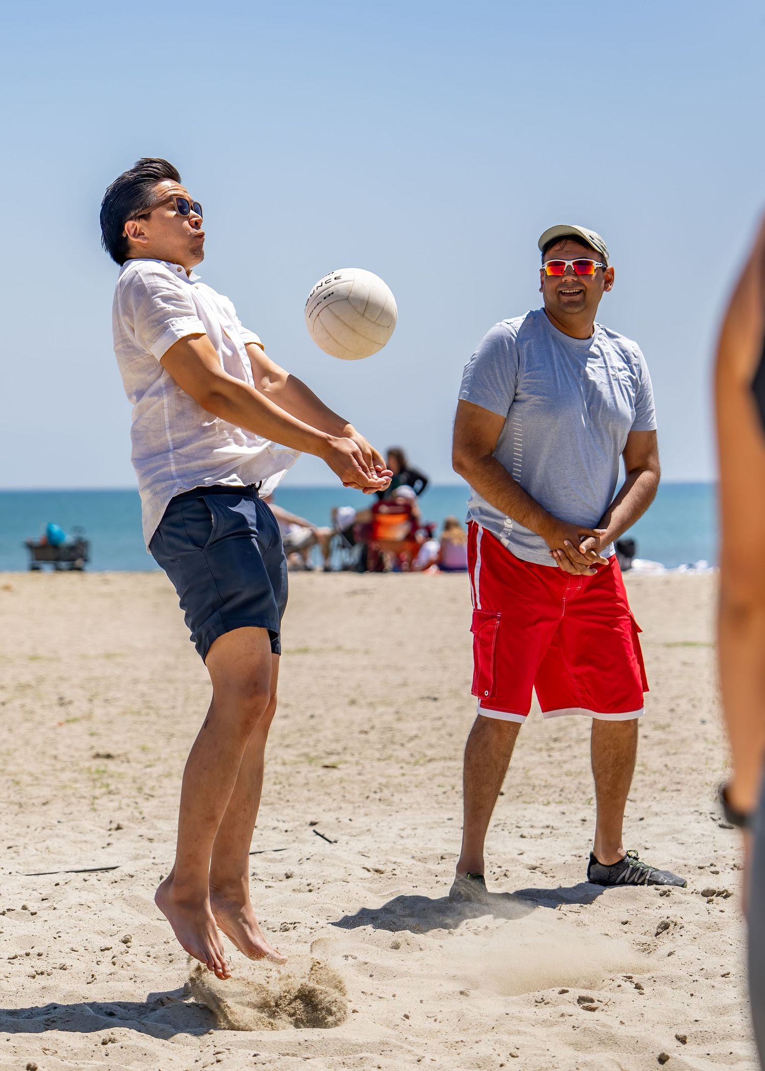 Two men playing beach volleyball, one jumping to hit the ball while the other watches nearby.