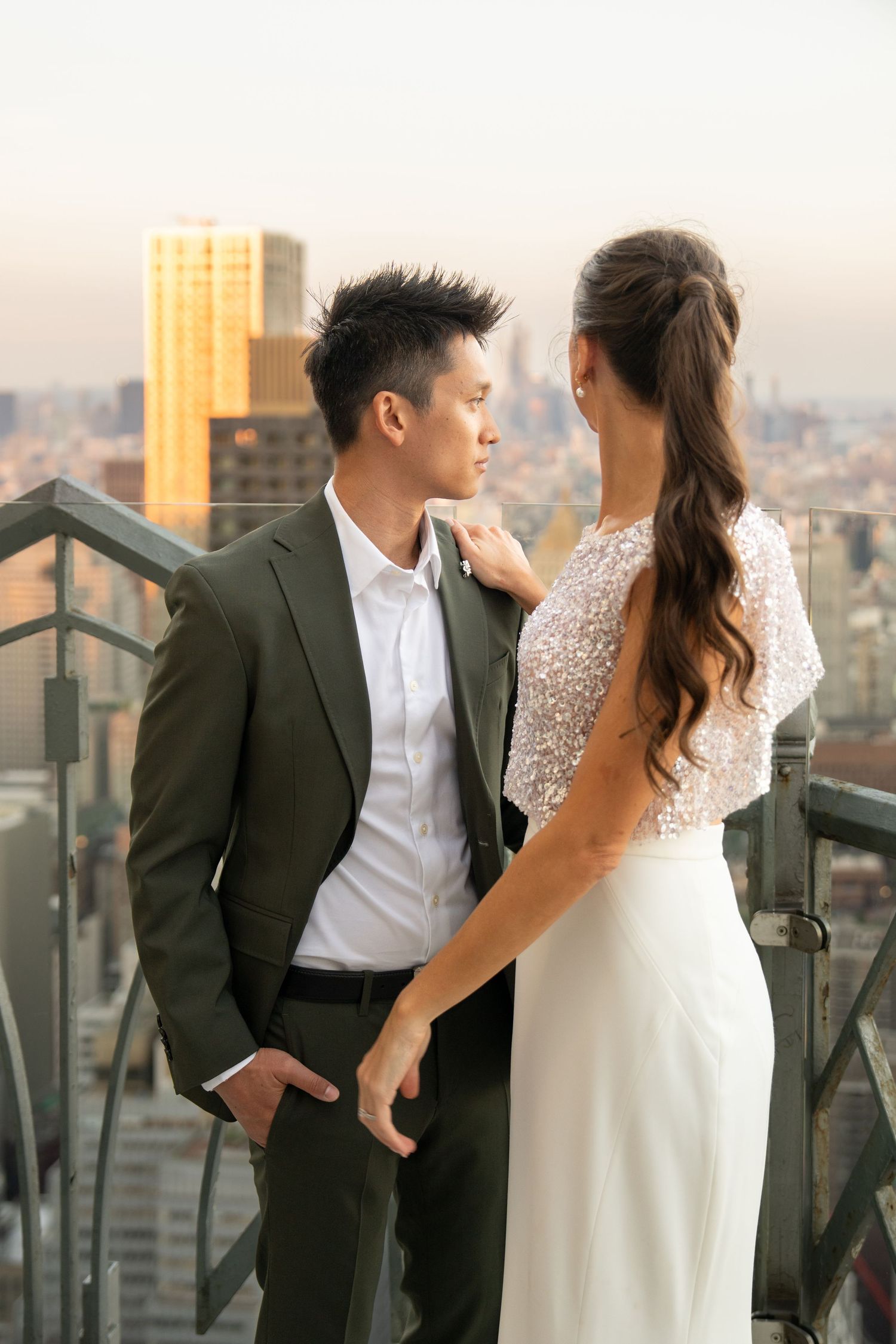 Couple in wedding attire sharing a romantic moment on a rooftop overlooking a city at sunset.