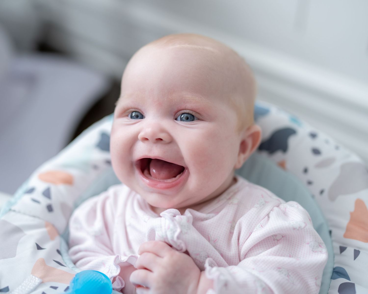 A cheerful blue-eyed baby in pink laughs while lying on a colorful blanket.