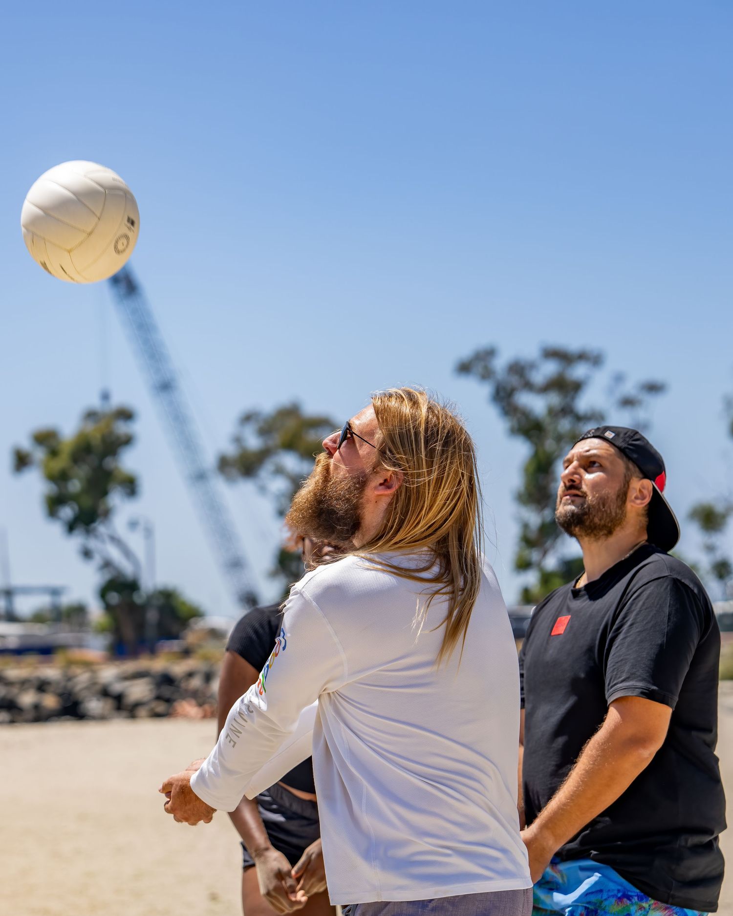 Two men watching a volleyball in the air on a sunny beach day.