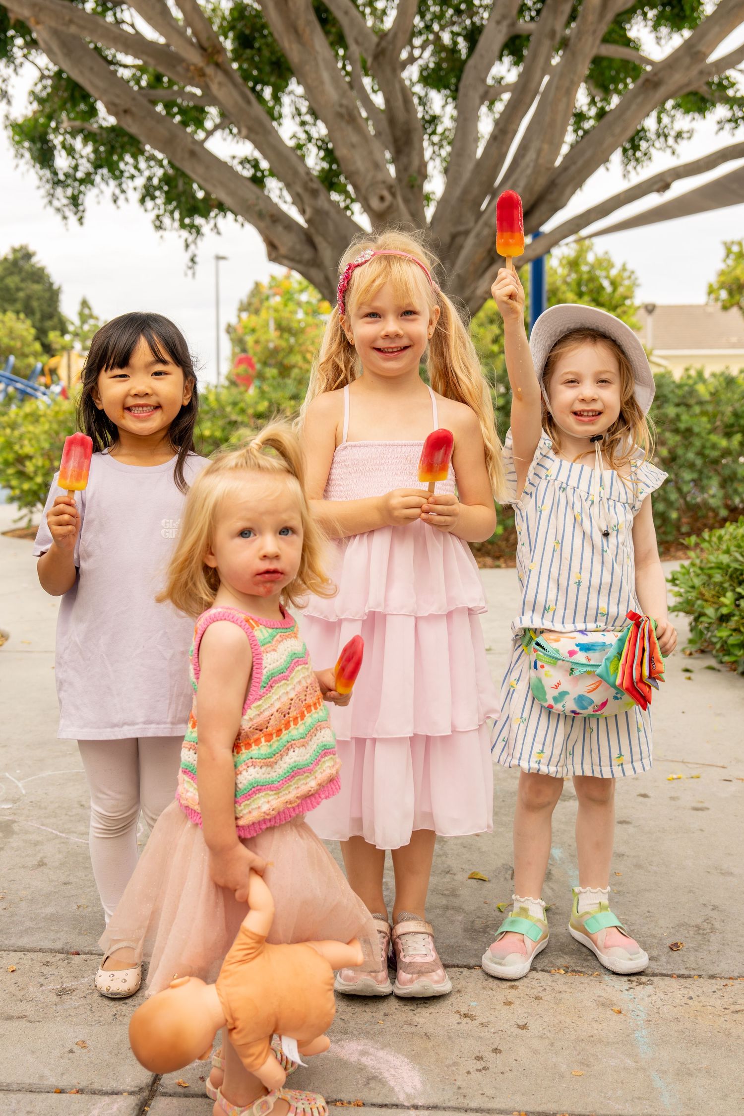 Four young girls holding colorful popsicles and smiling outdoors under a large tree.