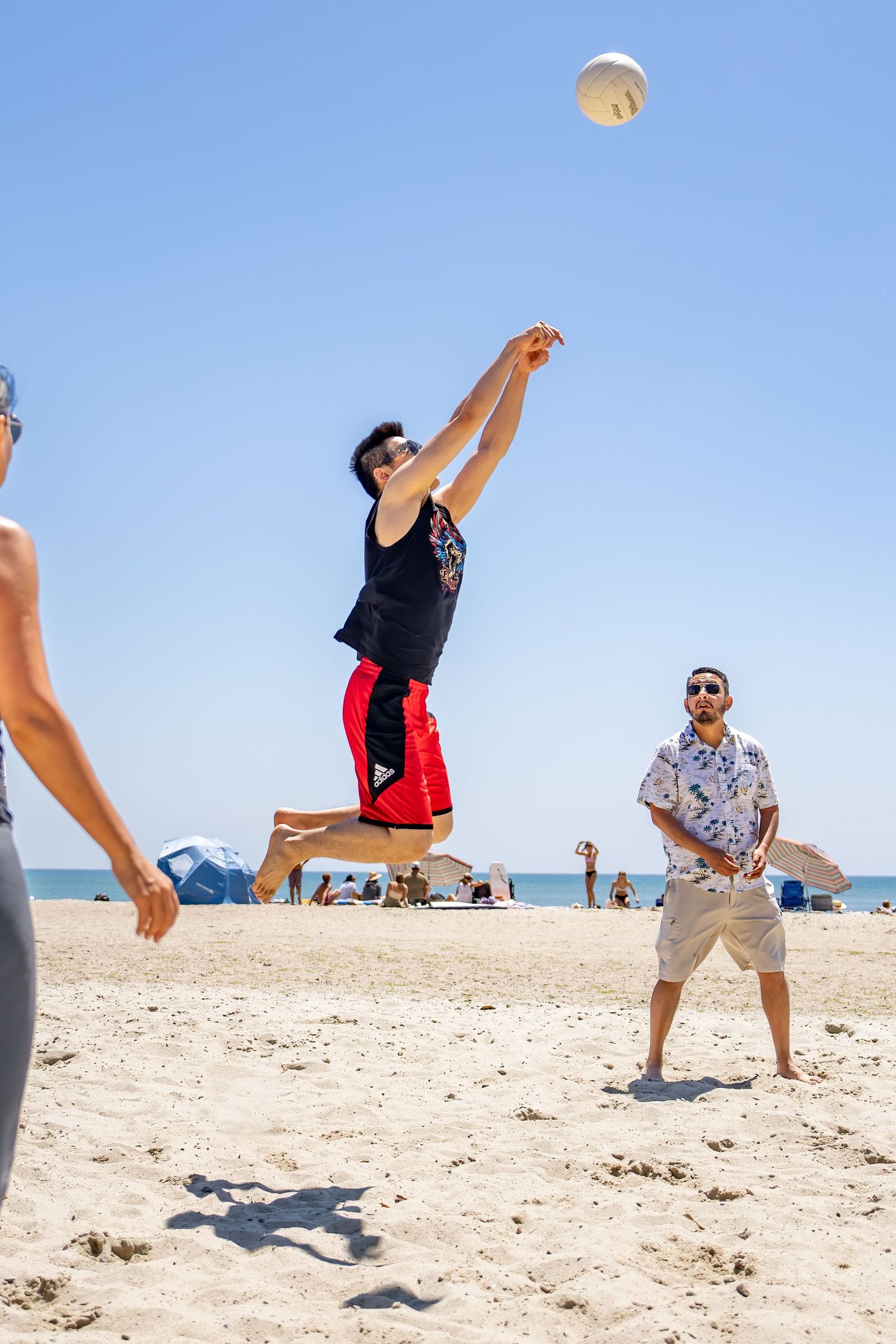 Man jumping to spike a volleyball on a crowded sunny beach with friends watching.