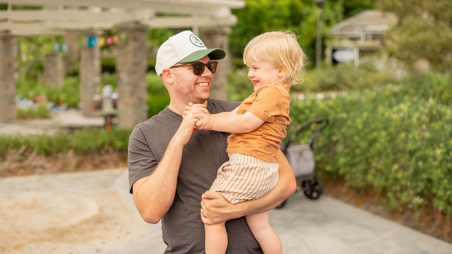 A smiling man in a white cap and sunglasses holds a happy toddler in an orange shirt outdoors.