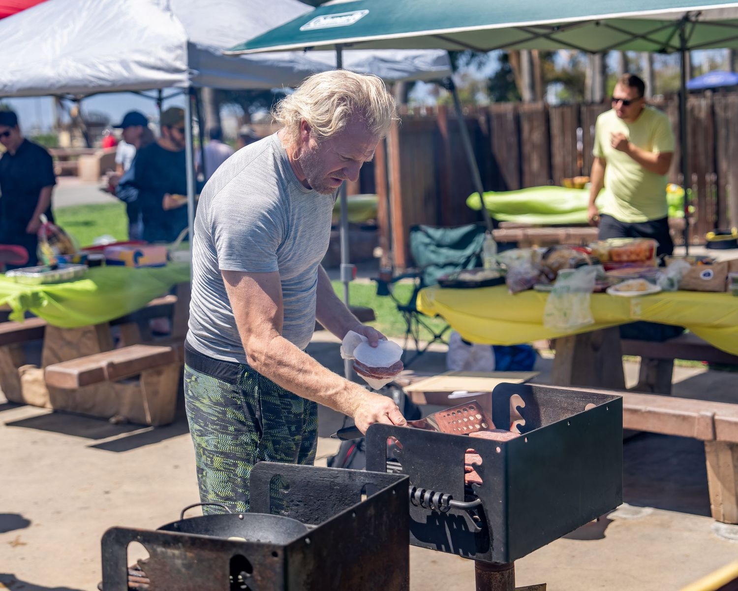 Older man with gray hair grilling meat on a charcoal BBQ grill at an outdoor beach event.