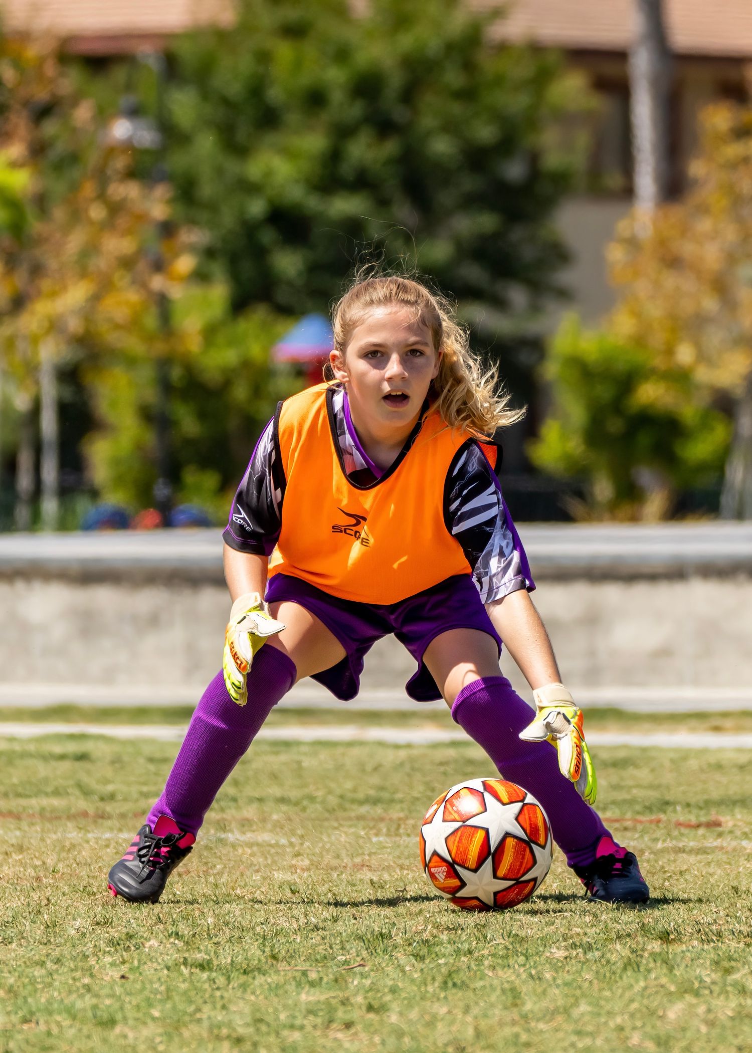 Girl goalkeeper in orange vest and purple socks ready to stop soccer ball on grass field.