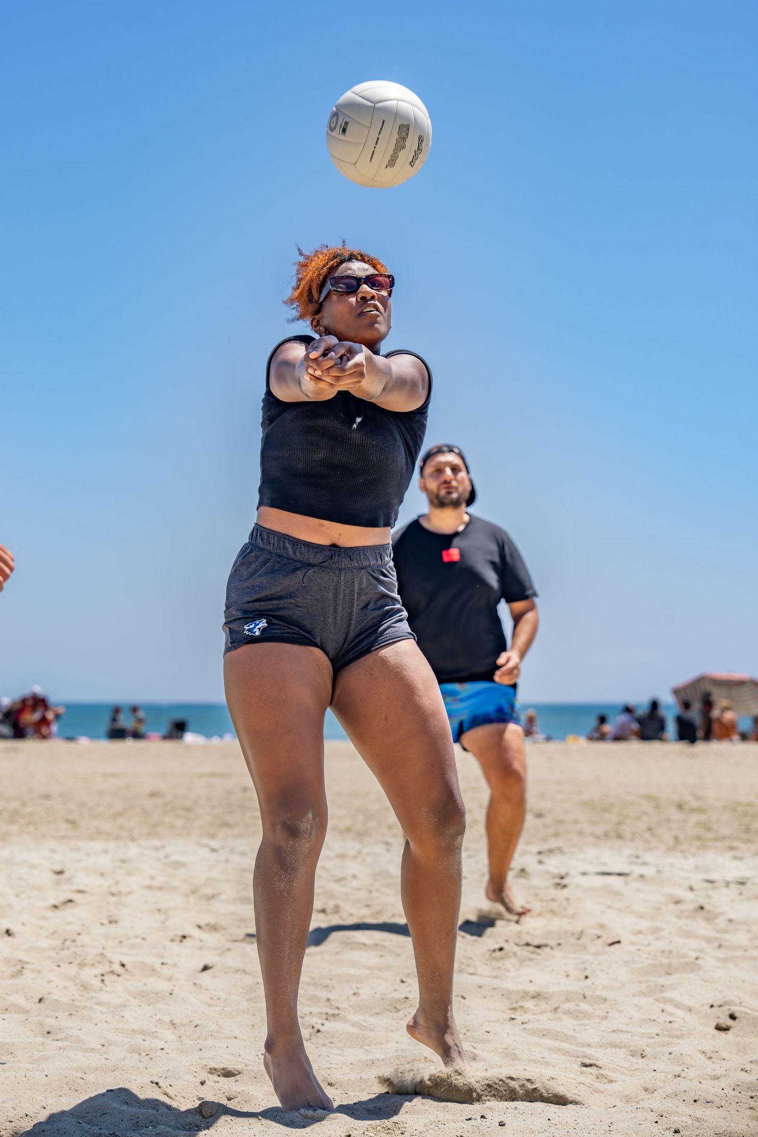 A woman bumps a volleyball on a sunny beach while a man watches in the background.
