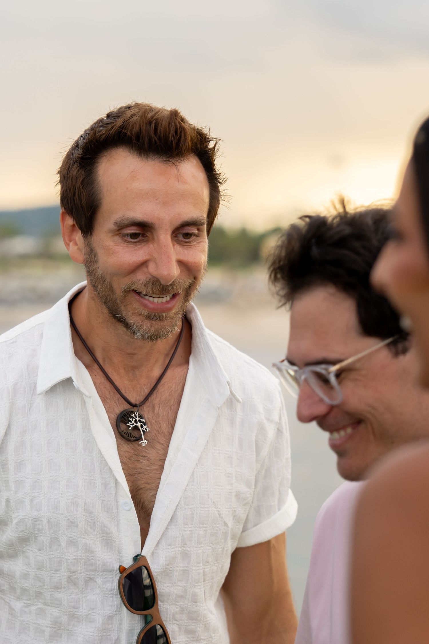 Smiling man in white linen shirt with necklace at a sunset beach gathering.