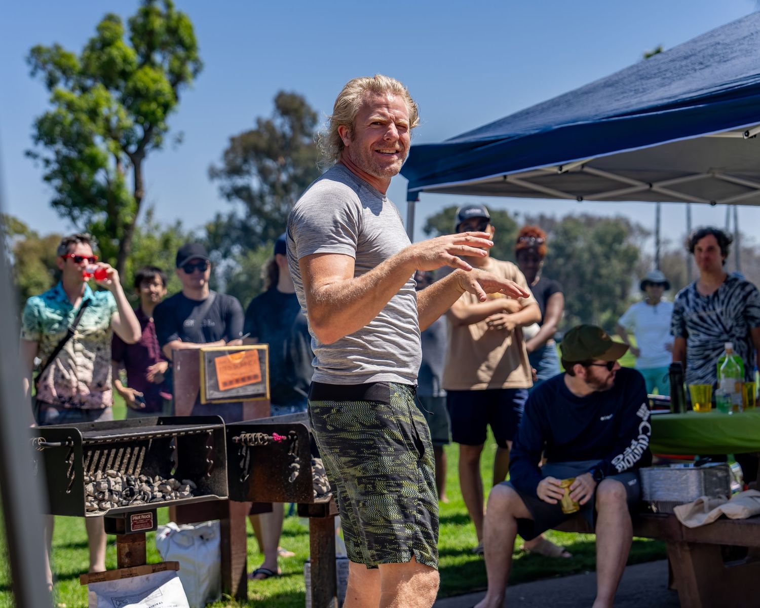 Fit older man in gray shirt holding a drink and smiling at an outdoor park gathering with people in background.