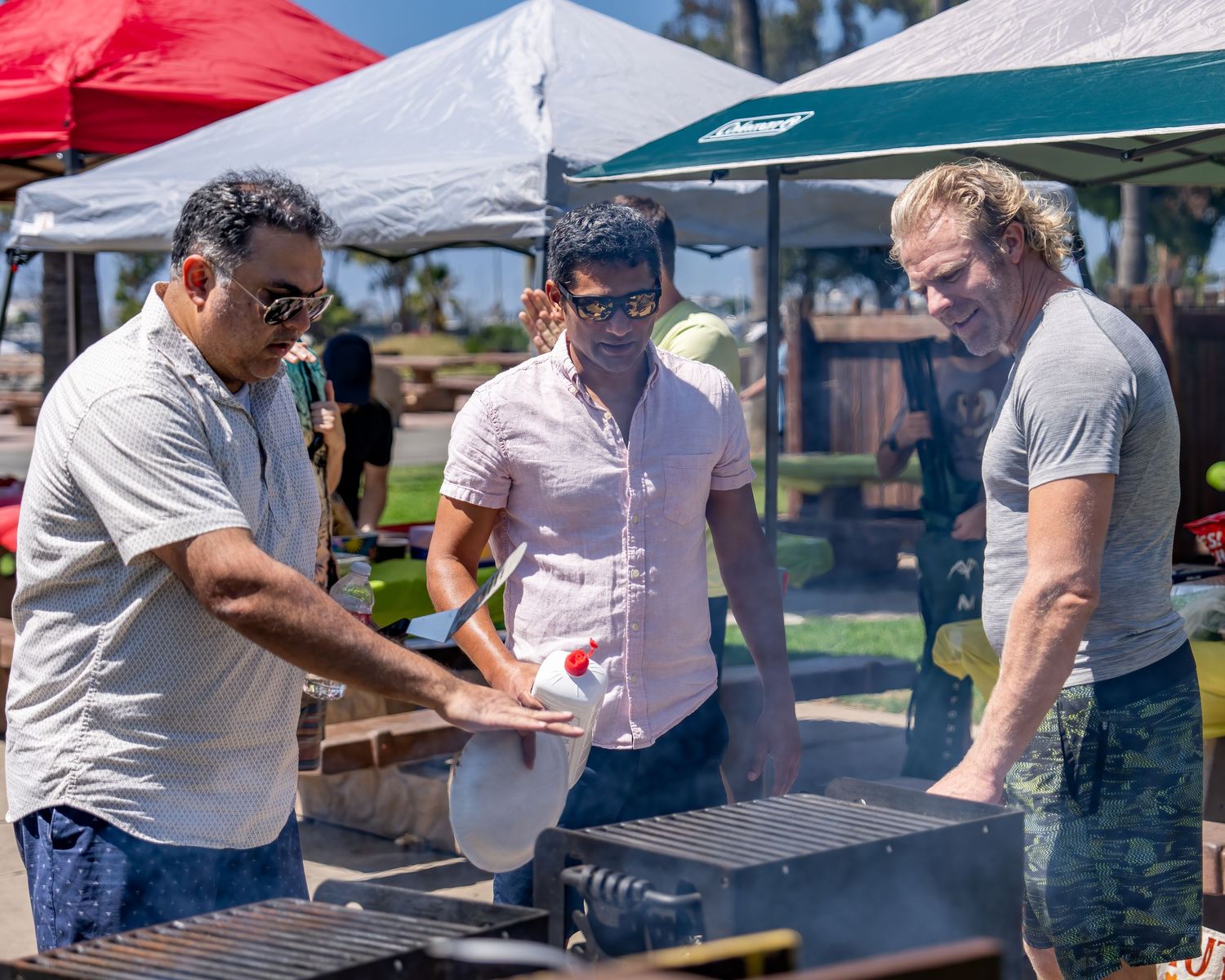 Three men working together around a smoking outdoor grill under canopy tents at a park.