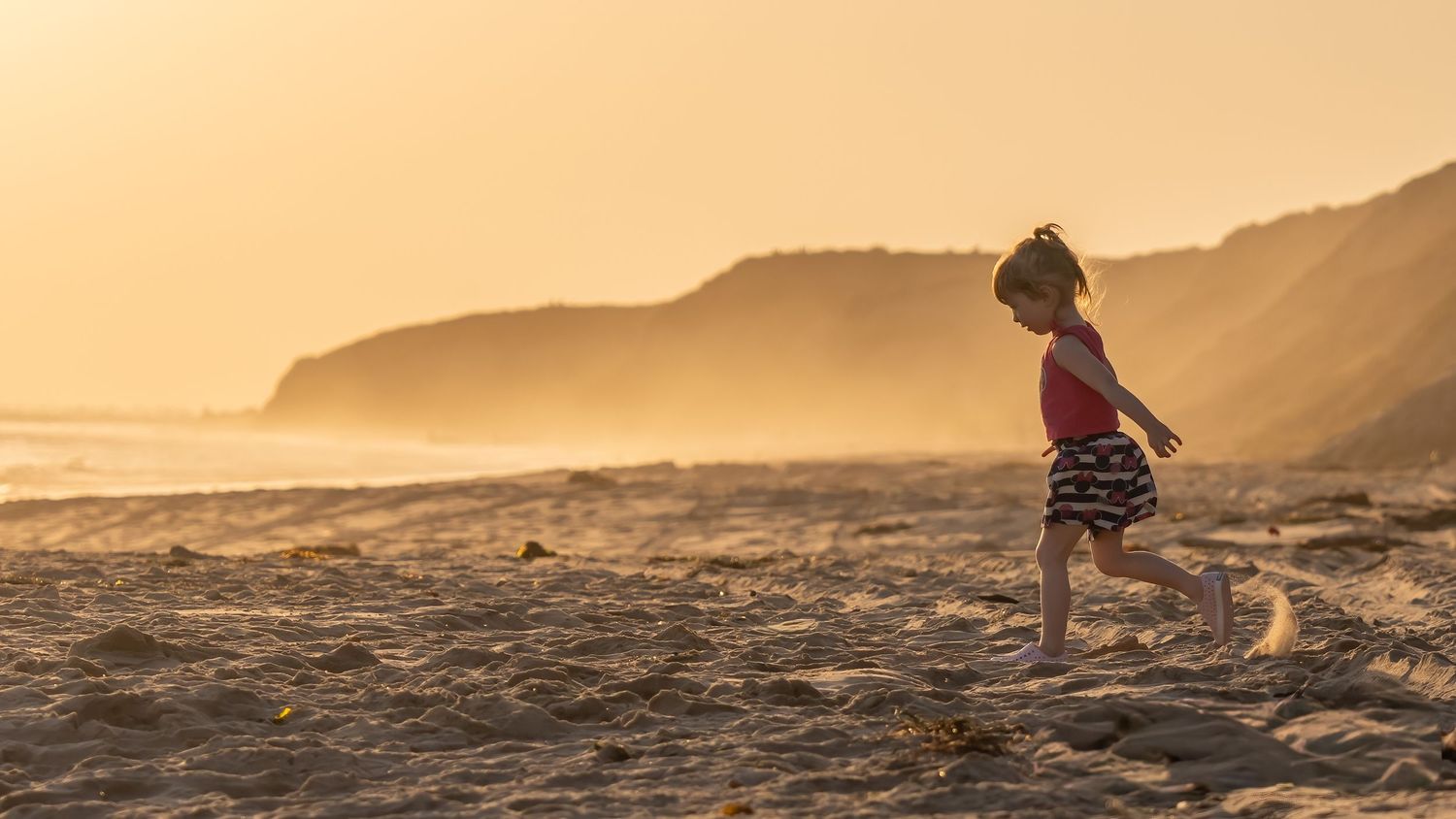 A young girl runs along a sandy beach at golden sunset with misty cliffs in the background.