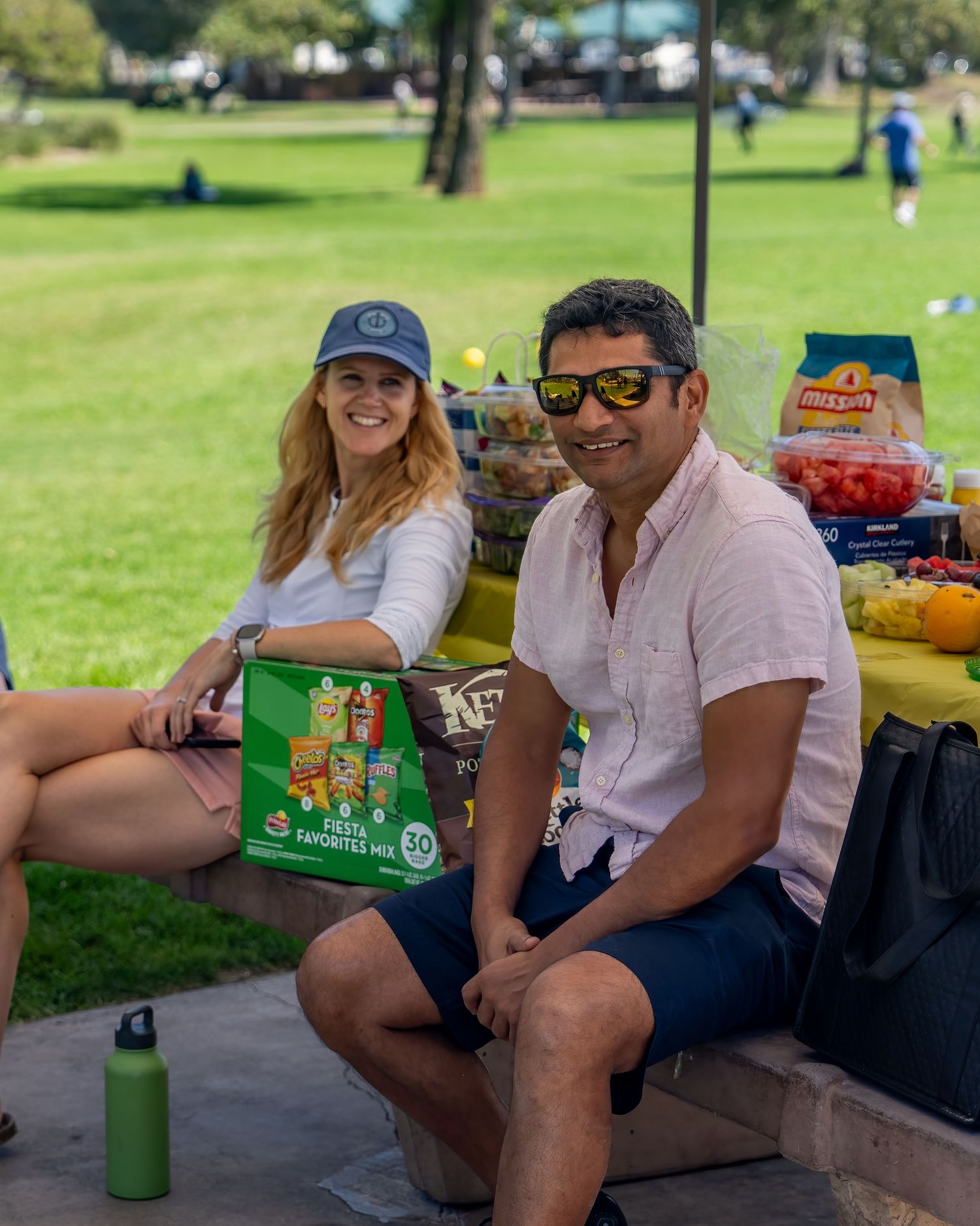 Smiling man and woman sitting at a picnic table with snacks and drinks at an outdoor event.