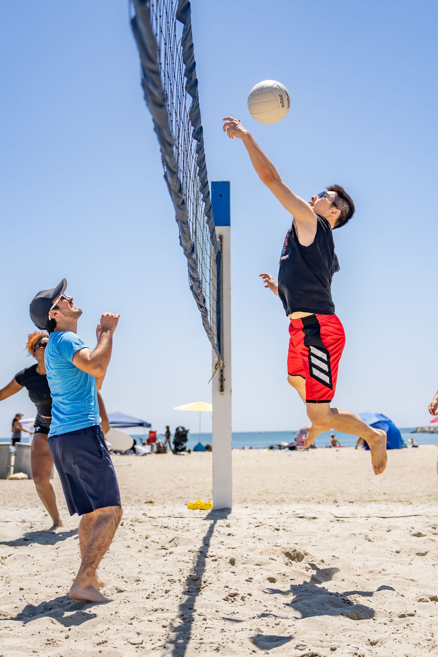 Young man jumps to spike volleyball over beach net on sunny day.