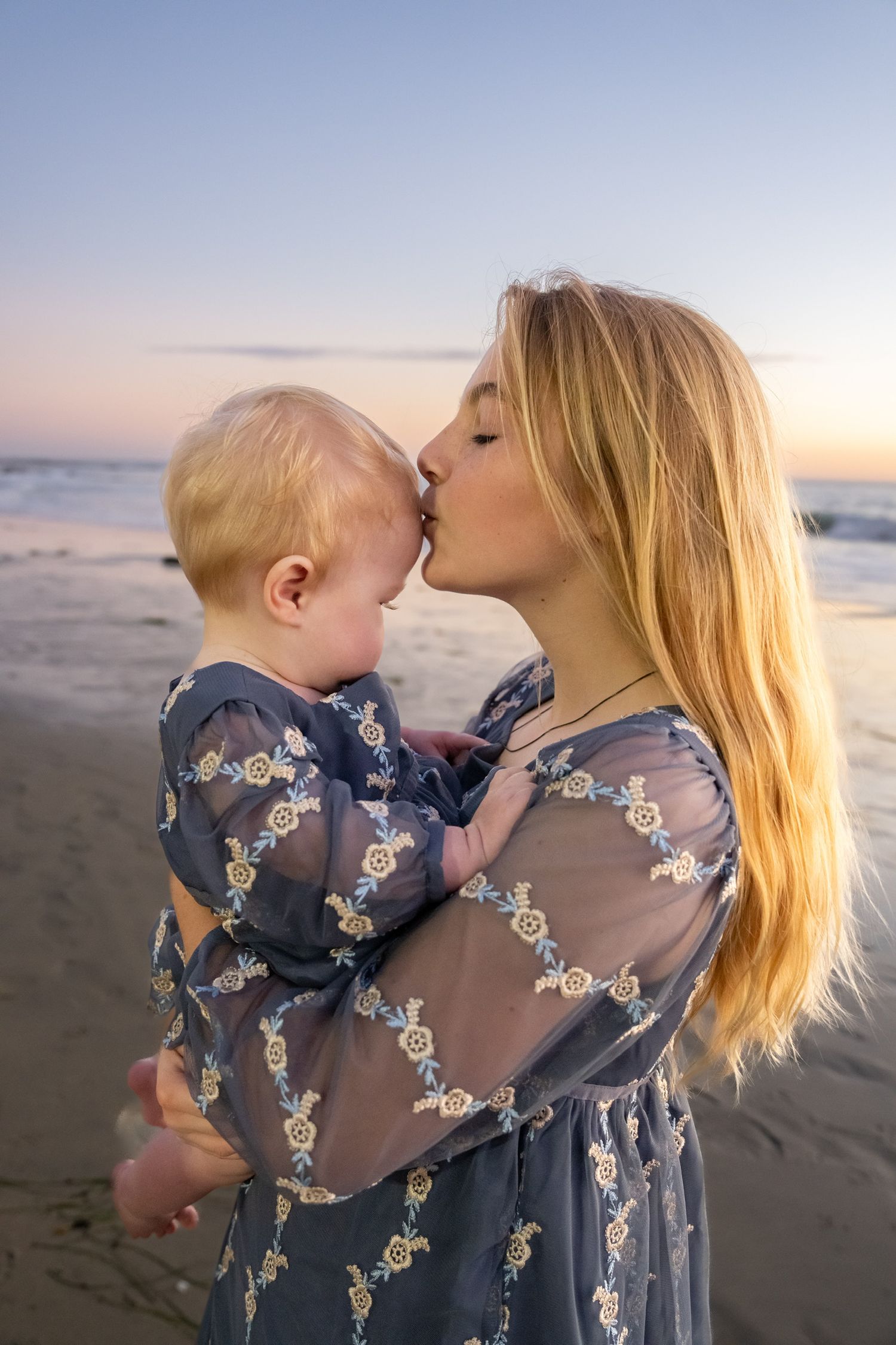 A blonde woman kisses a baby on the cheek at a beach during a beautiful pastel sunset.