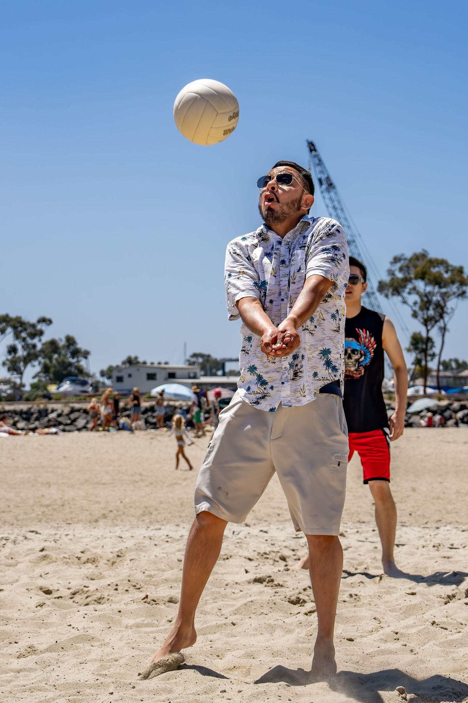 Man in floral shirt bumping volleyball on sandy beach on sunny day.