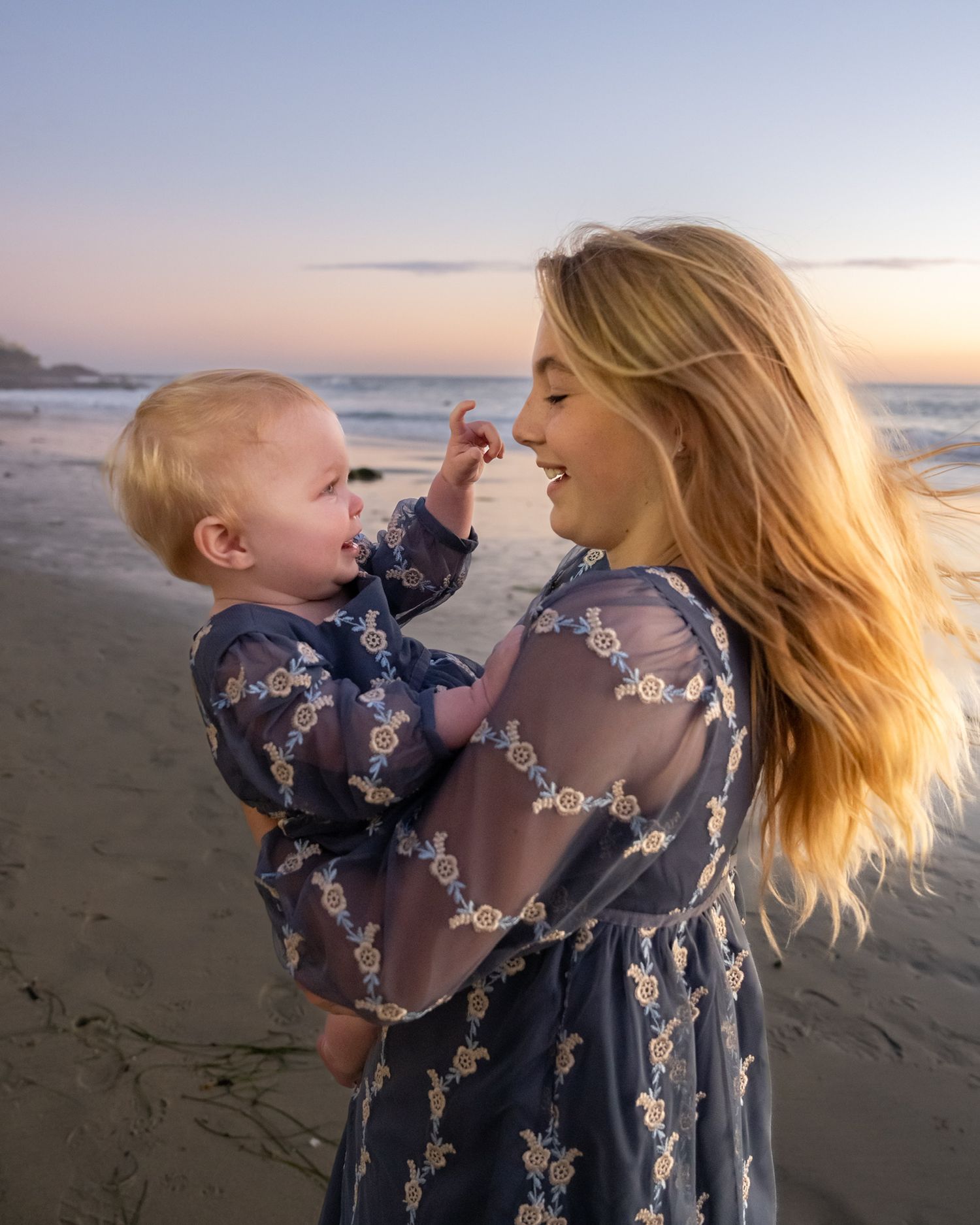A smiling blonde woman holds a baby on a beach at sunset, both wearing matching blue floral dresses.