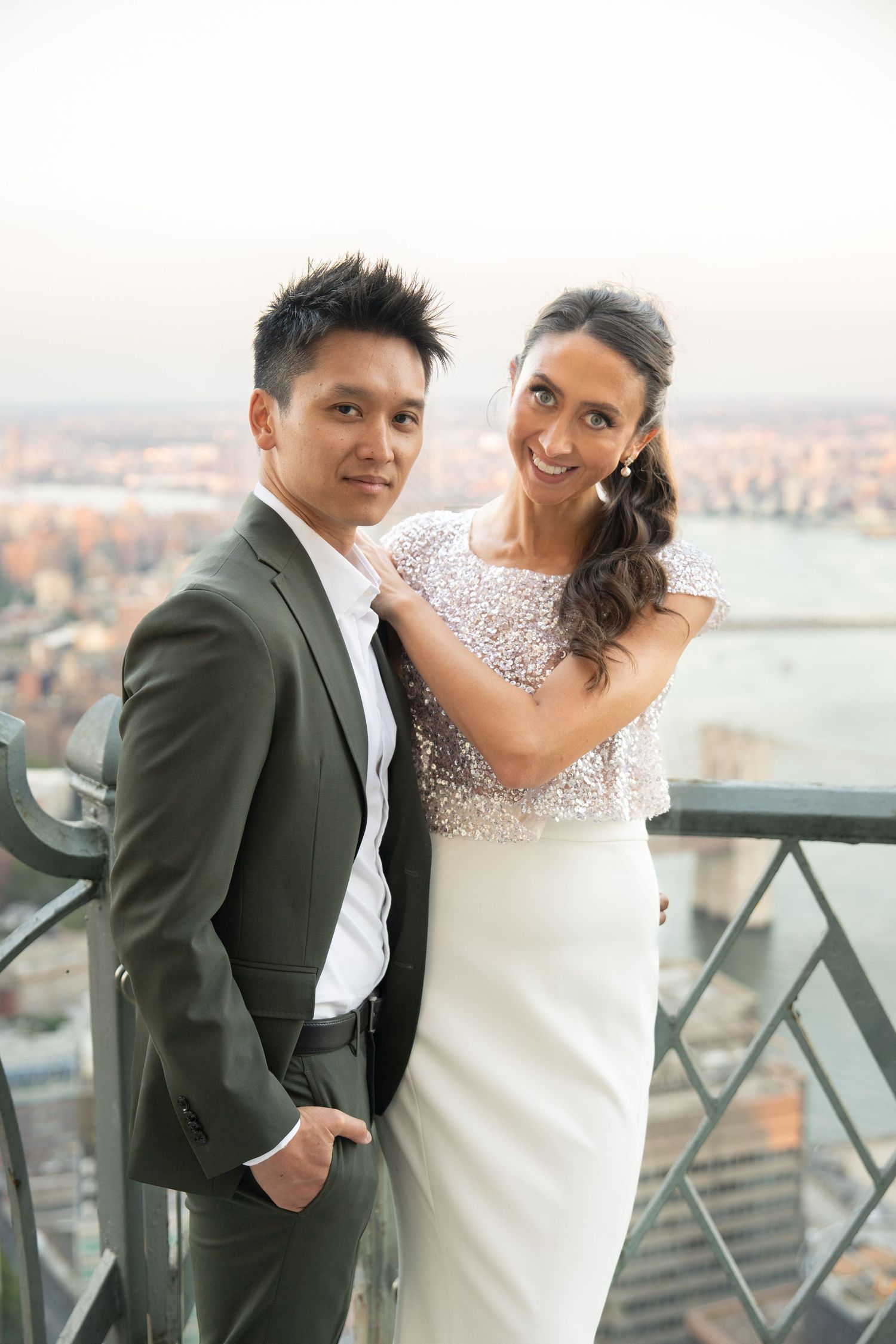 Elegant couple in wedding attire posing on a rooftop with a city skyline at sunset.