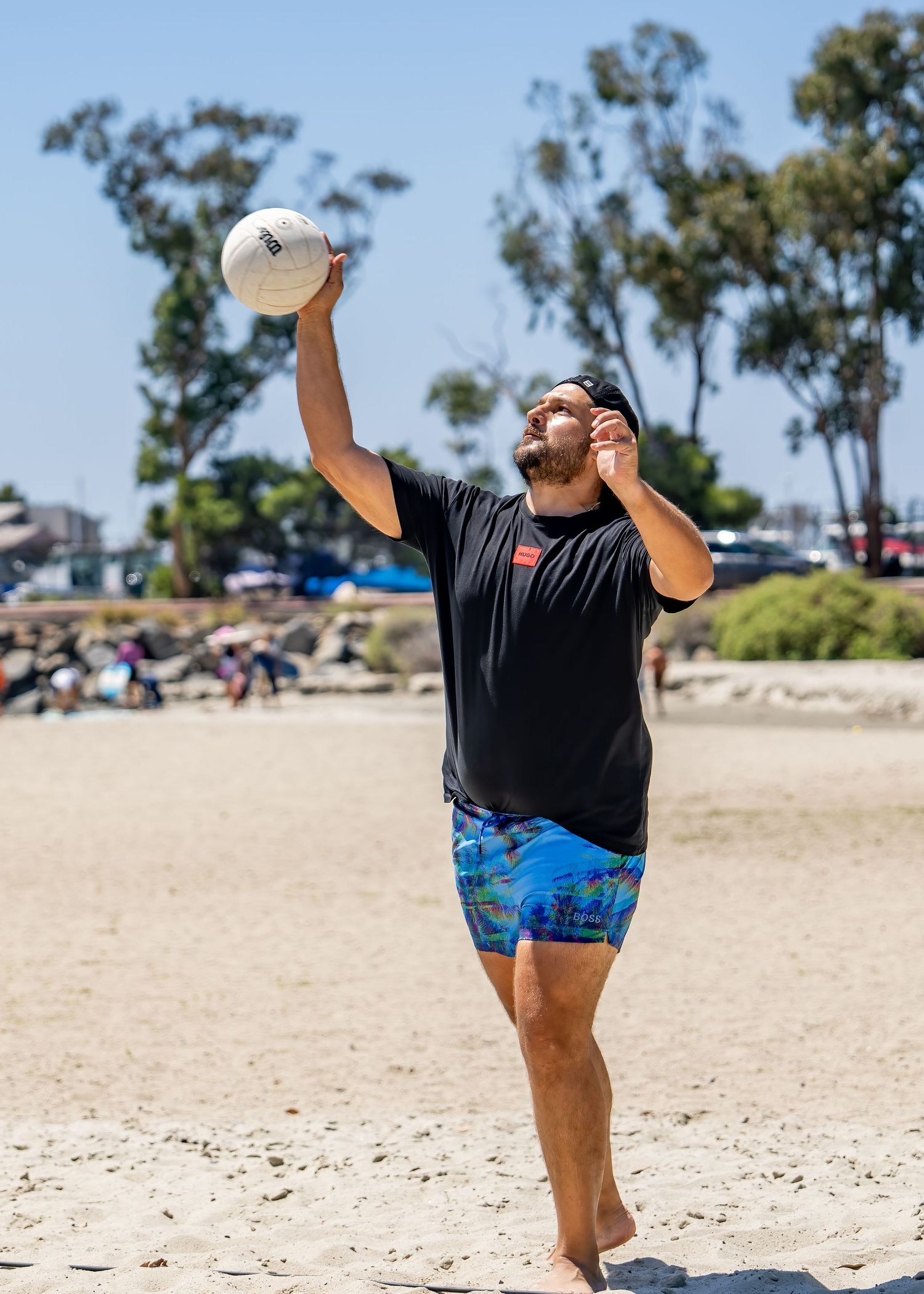 Man in black shirt and blue shorts serving a volleyball on a sunny beach with trees in background.