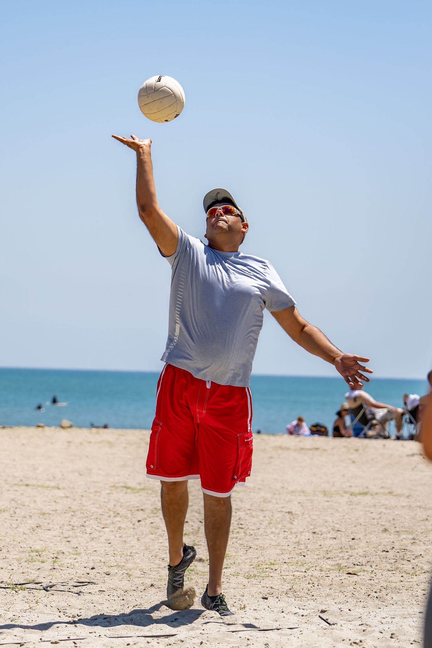 Man in red shorts and white shirt serving a volleyball on a sunny beach with ocean behind him.