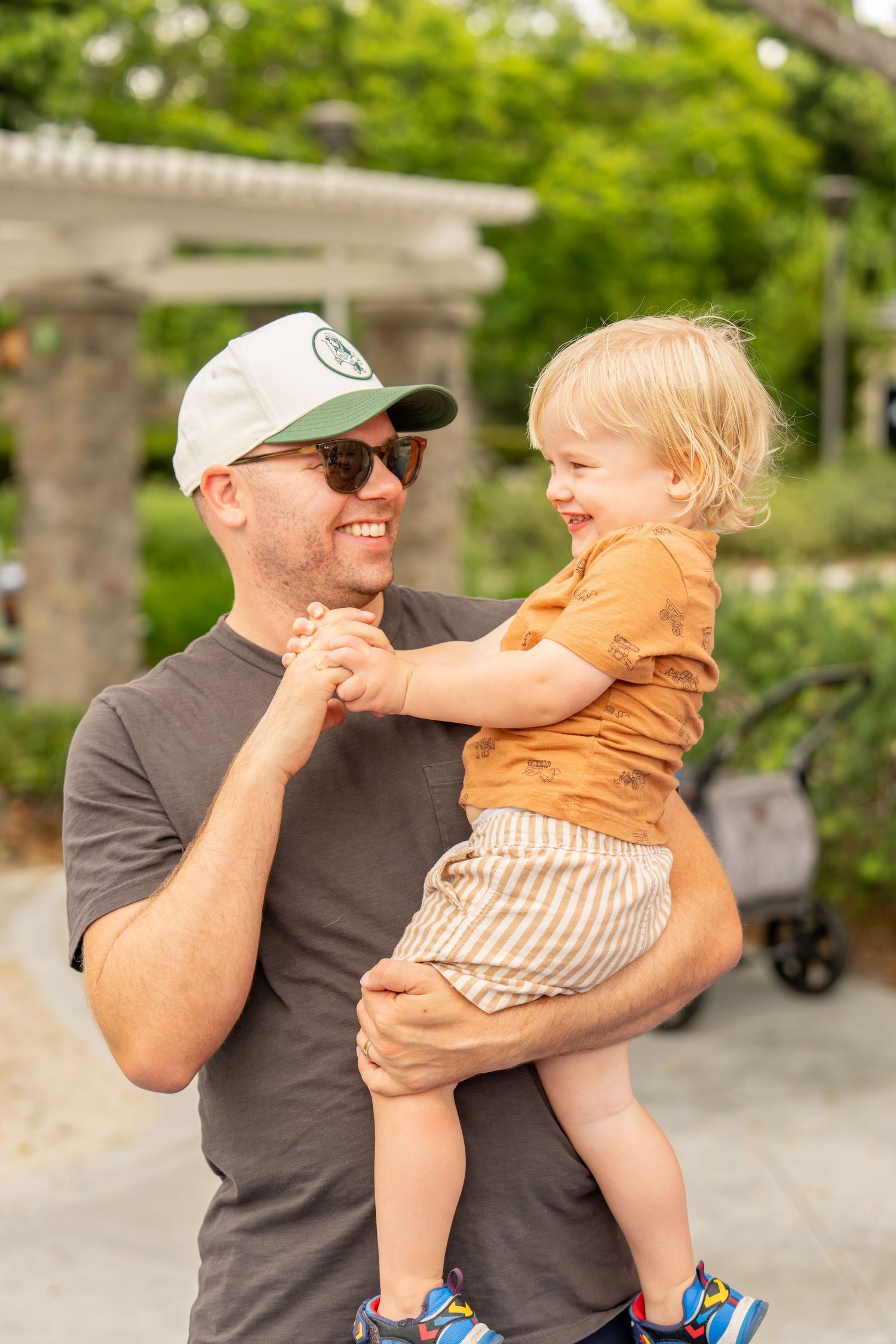 A smiling man in a cap holds a laughing toddler outdoors near greenery.